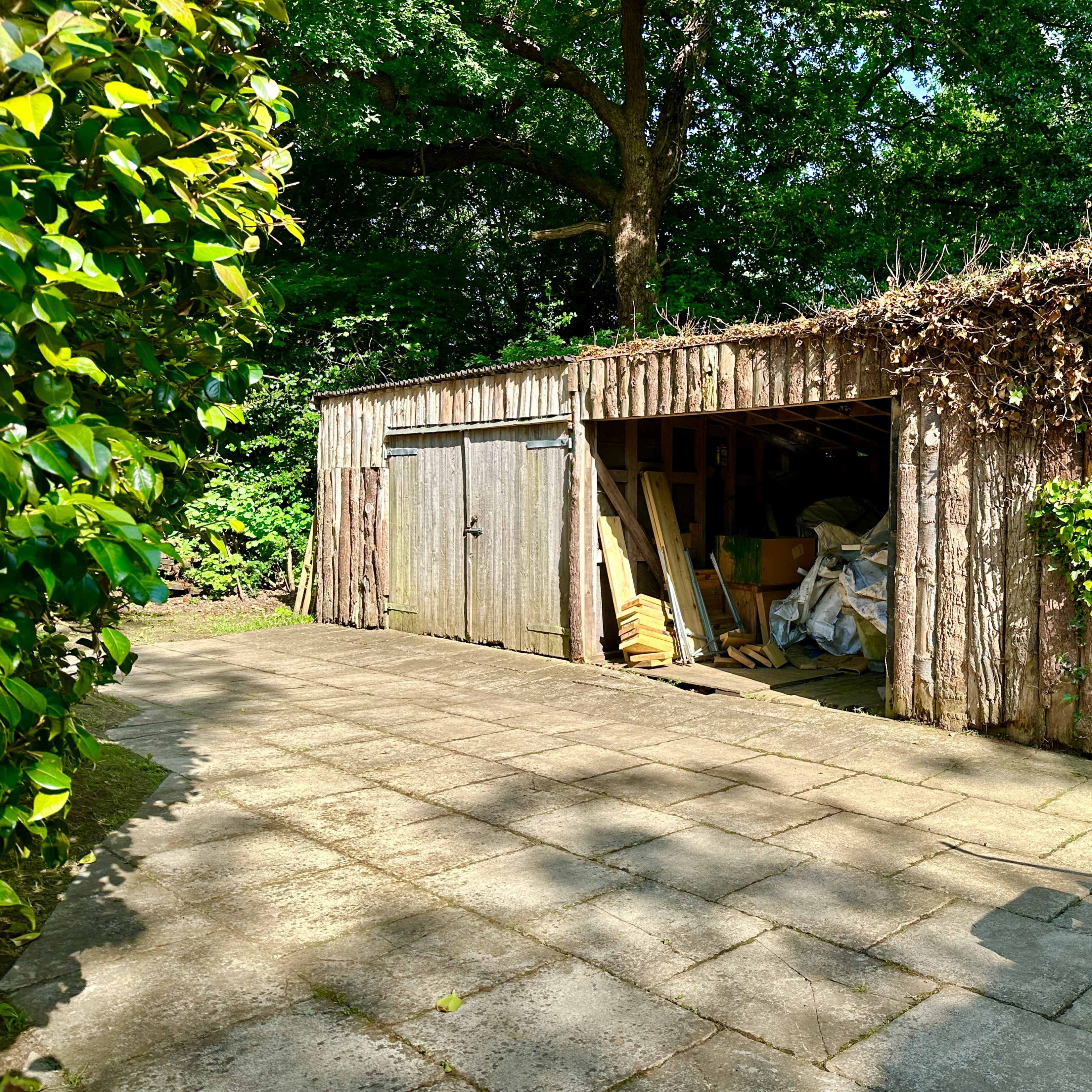 A wooden shed with overgrown vines is set on a stone patio surrounded by greenery.
