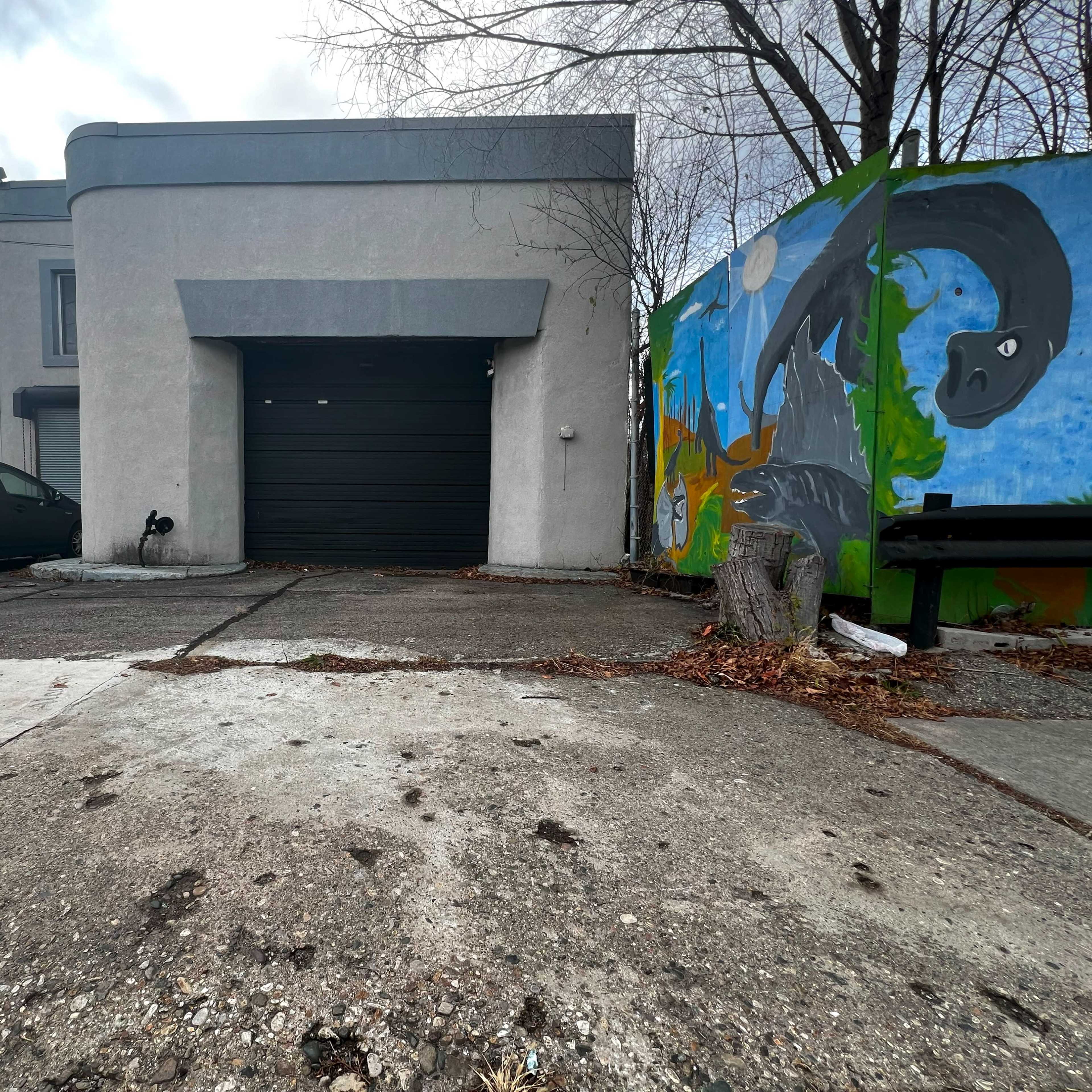 A gray building with a black garage door is adjacent to a colorful mural and a parked car, set against a cloudy sky.