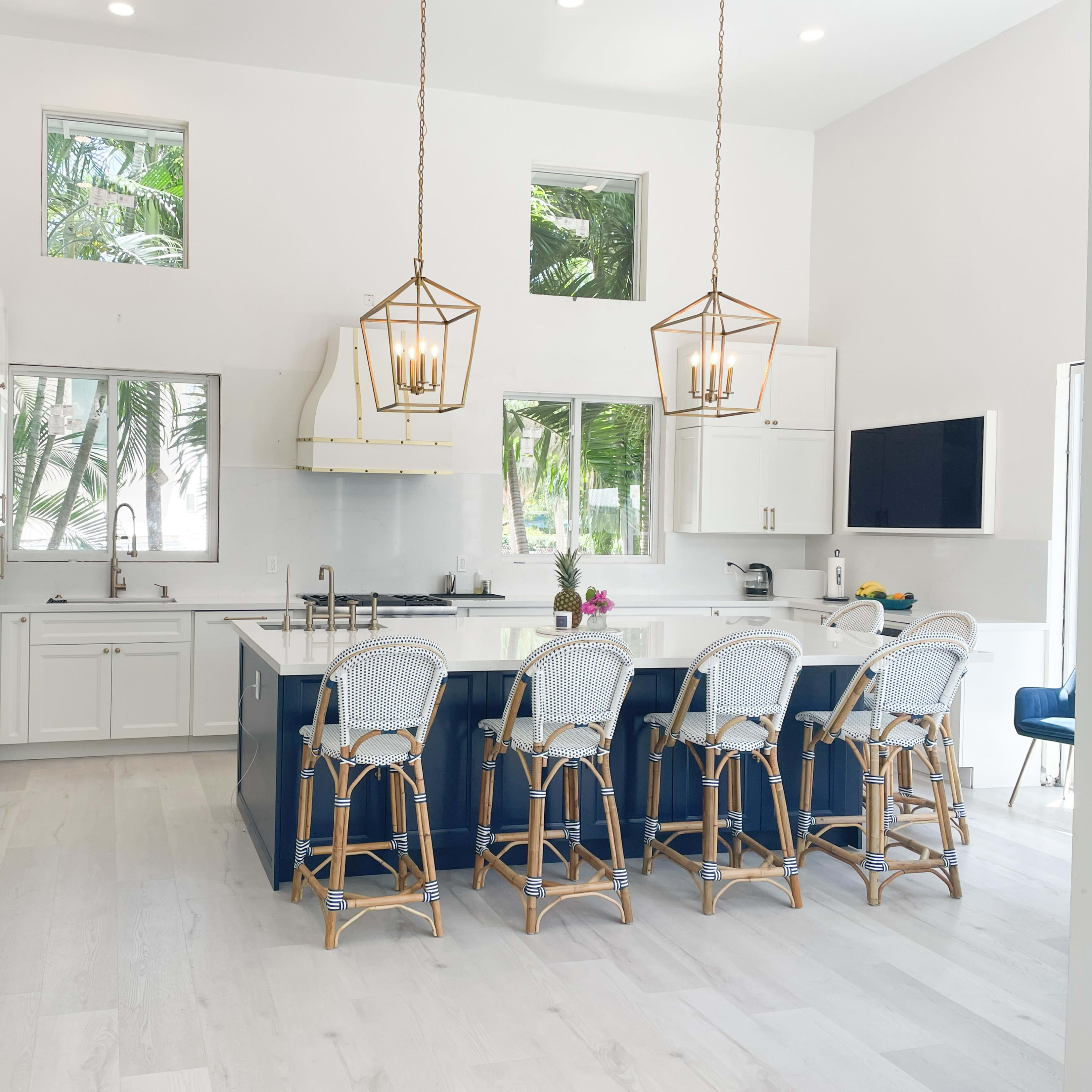 A modern kitchen features a large island with six barstools, white cabinets, stainless steel appliances, and large windows overlooking tropical greenery.