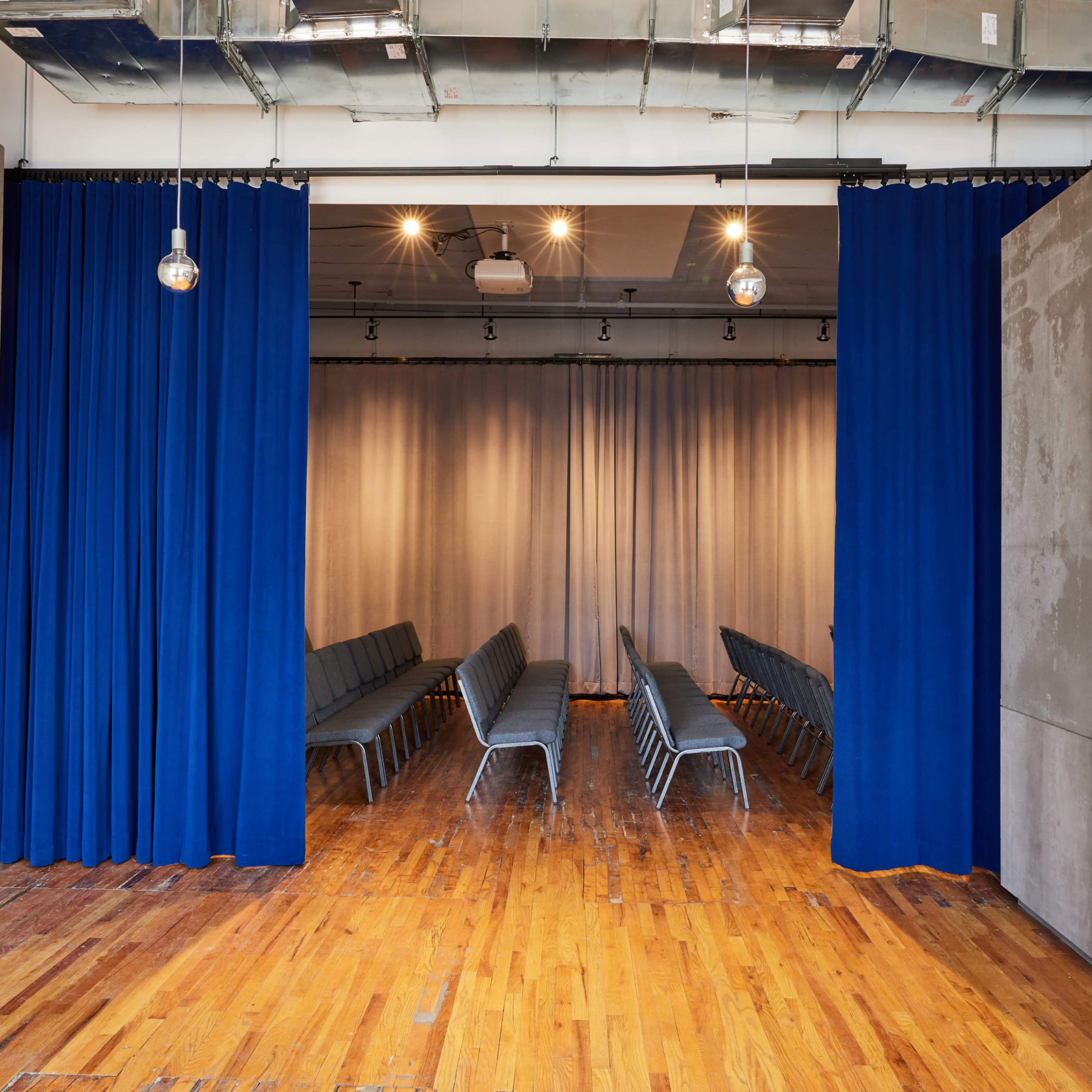 A conference room with rows of gray chairs facing a curtain-draped stage, flanked by blue curtains and walls of exposed concrete.