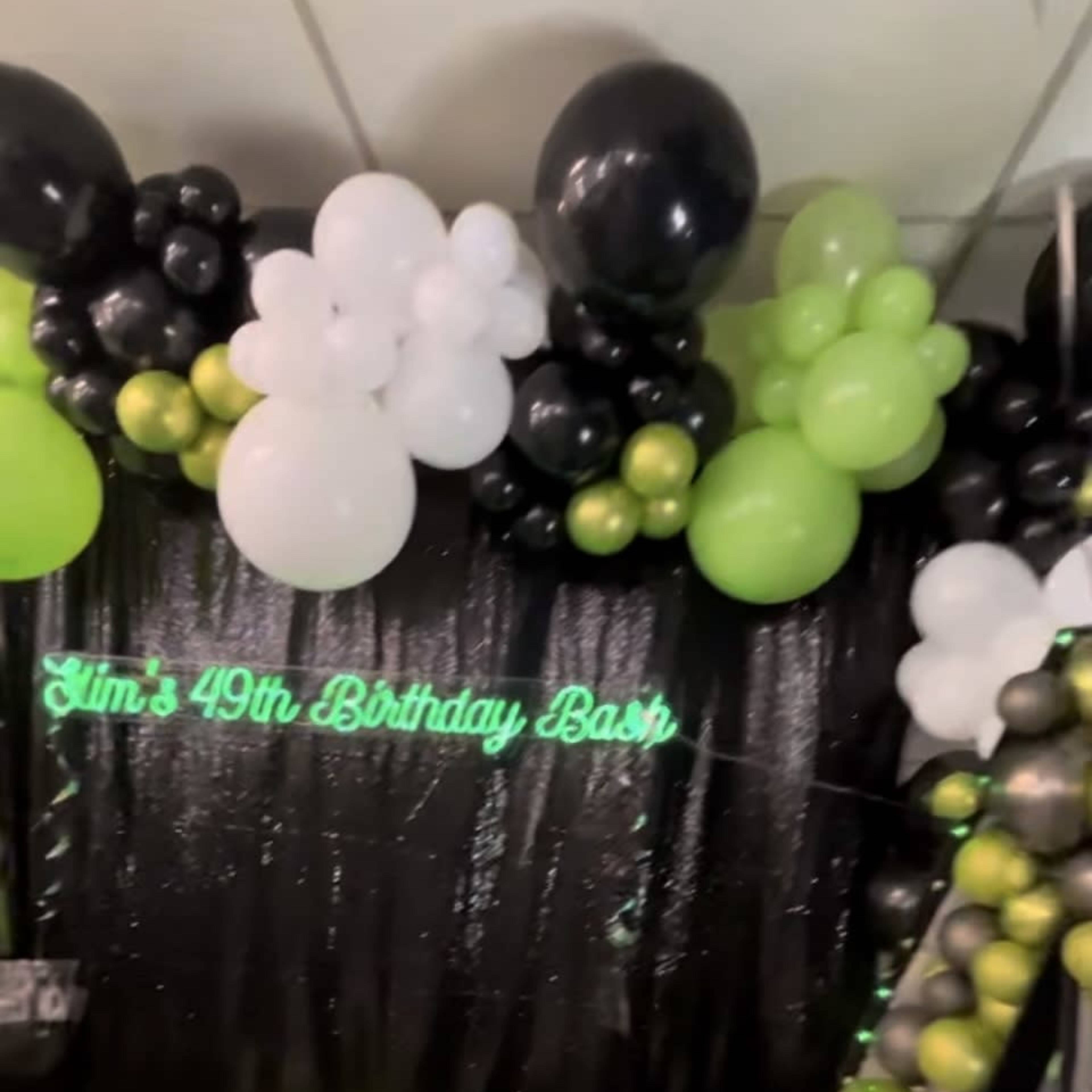 A black and green balloon arch with a sign that reads "Jim's 49th Birthday Bash" against a shimmering background.