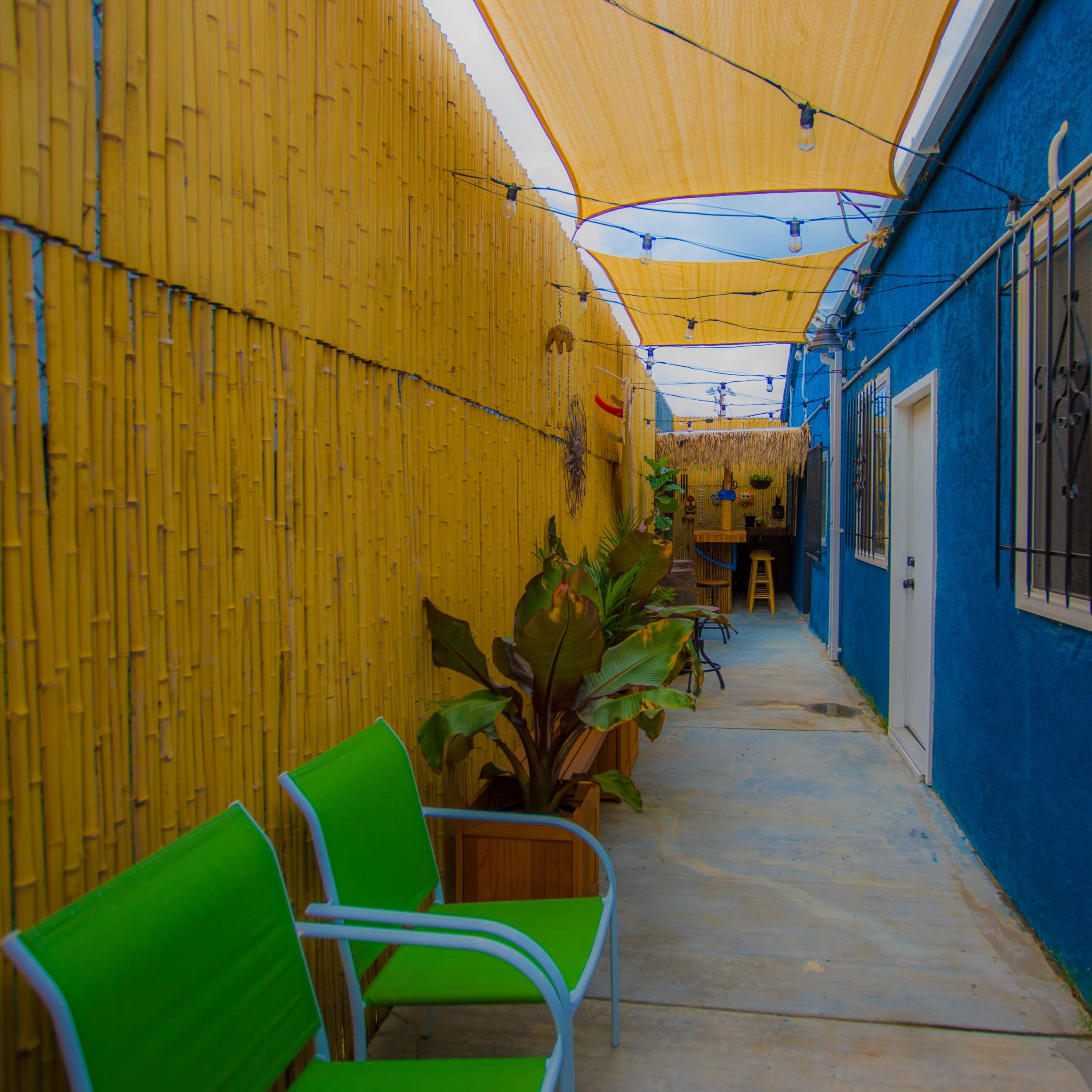 A narrow outdoor corridor is lined with yellow bamboo walls and features green chairs, leading to a brightly painted blue building.