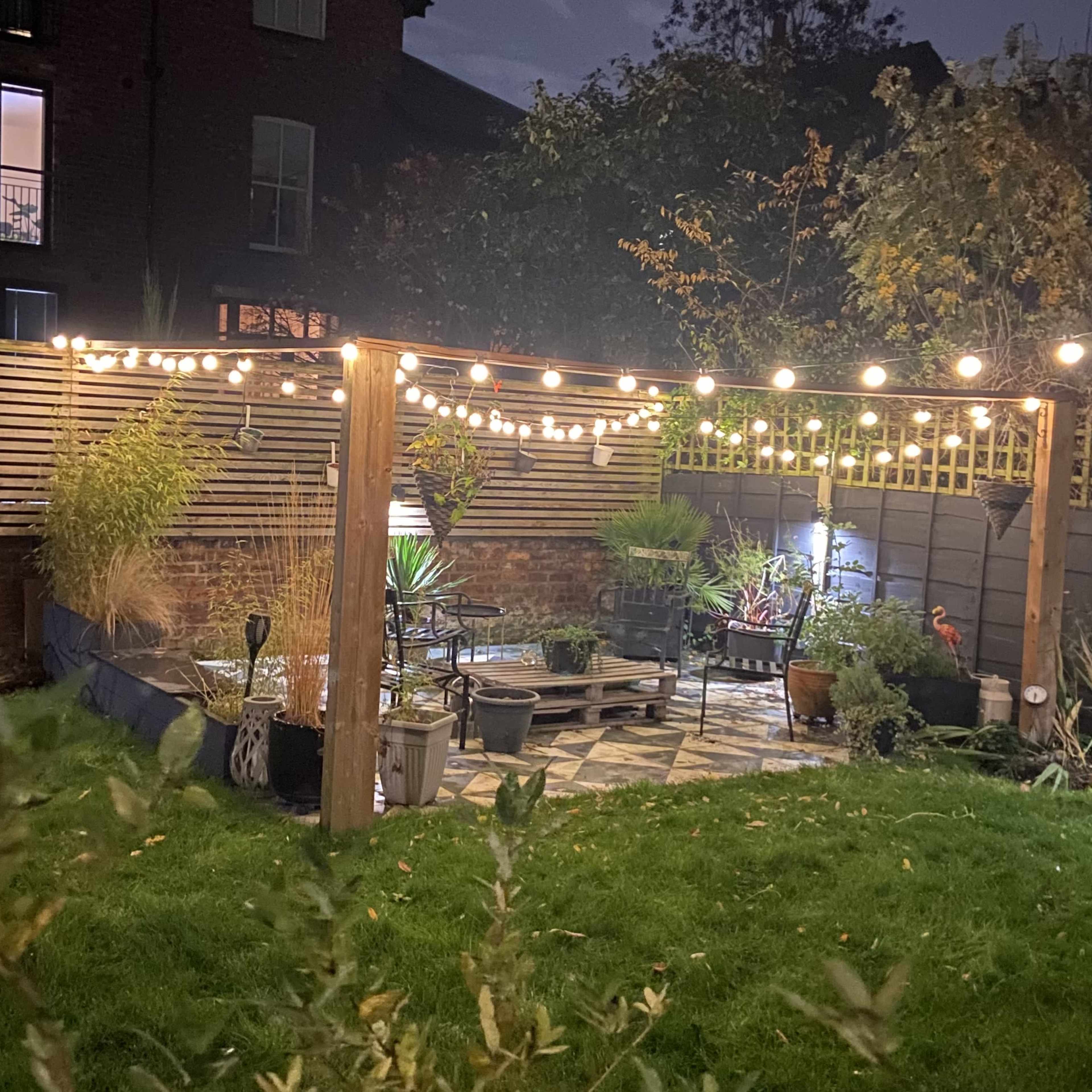A garden at night with a wooden pergola adorned with string lights, surrounded by various plants and seating areas on a stone patio.