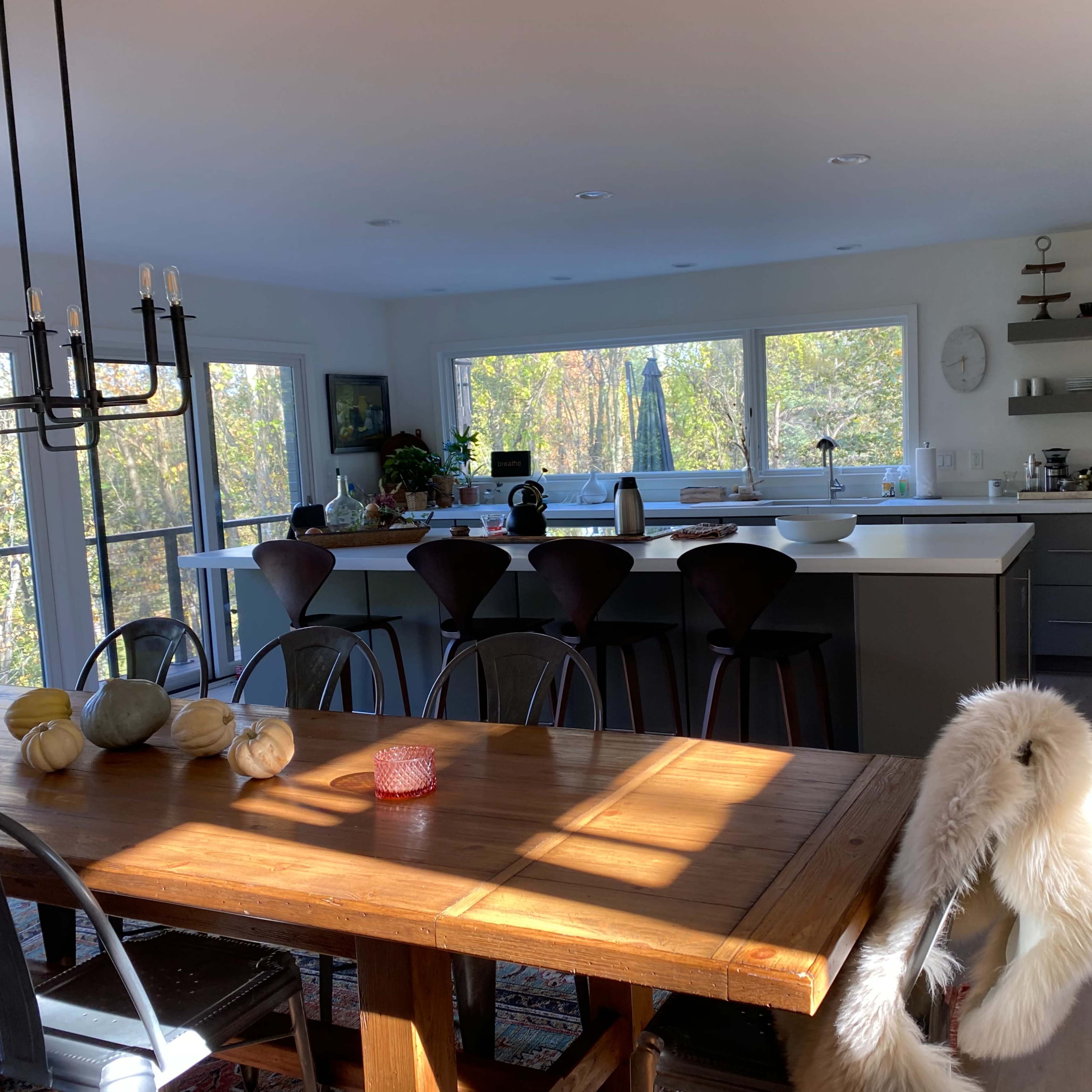 A modern kitchen with a large island and a dining table, featuring large windows that offer a view of greenery outside.