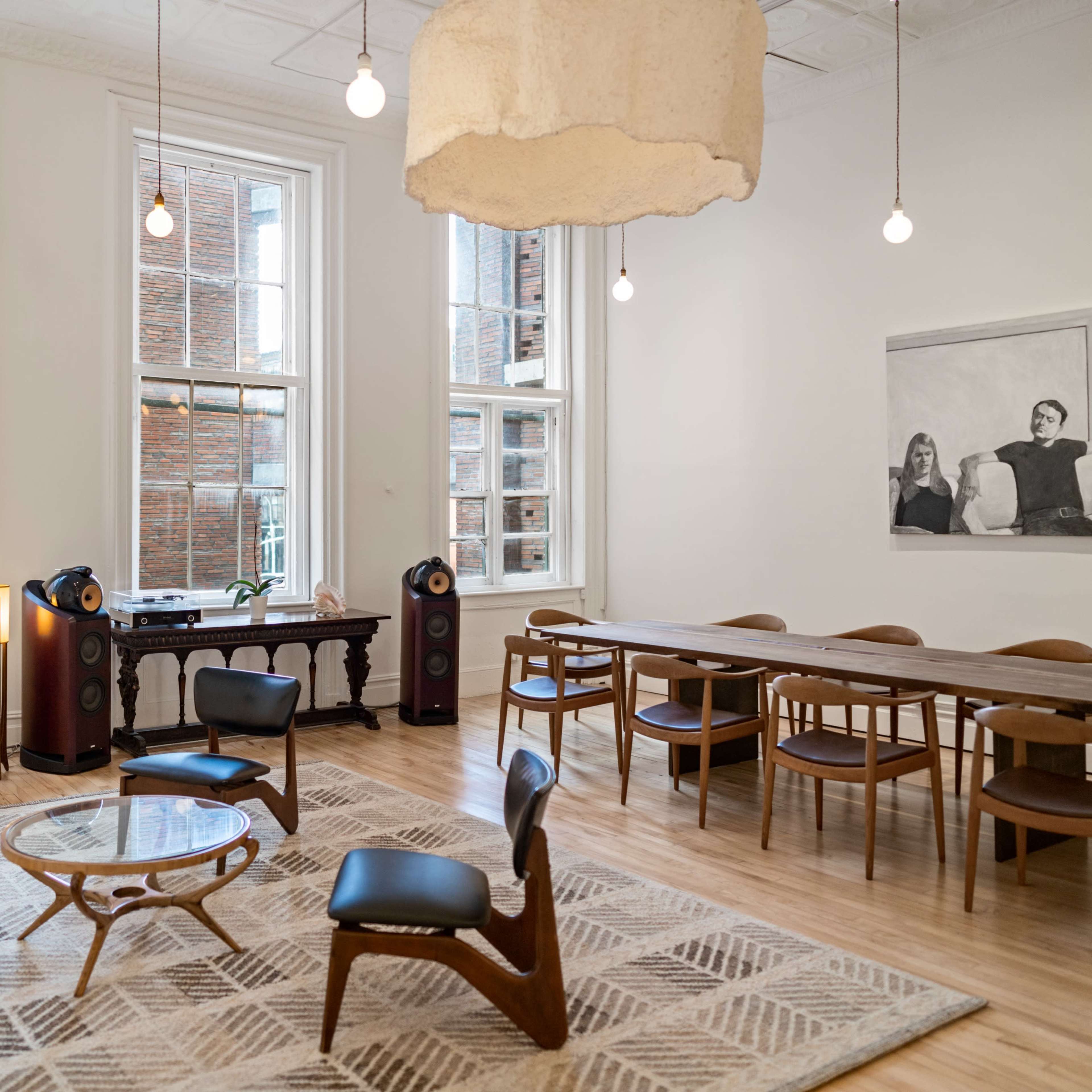 A spacious room with large windows, wooden floors, a dining table, and modern furniture including a lounge chair and audio equipment.