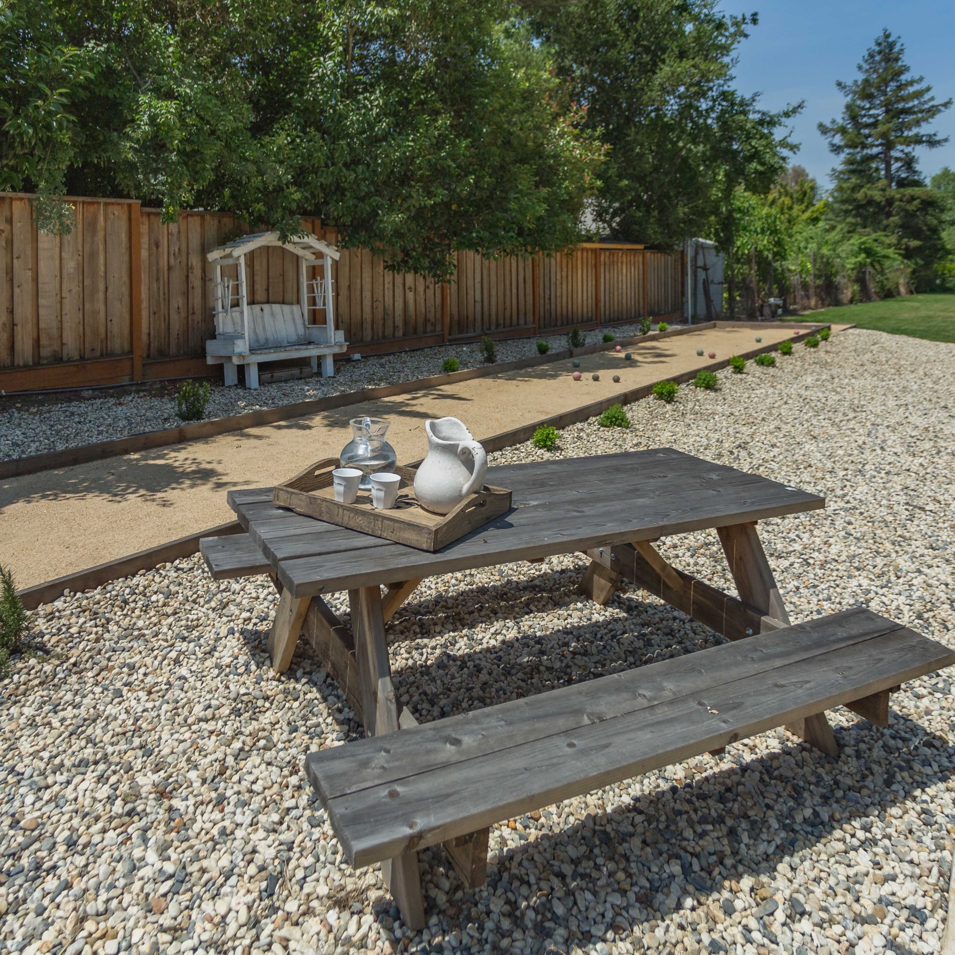 A wooden picnic table sits on gravel beside a landscaped area with a bocce ball court and a seating structure in the background.