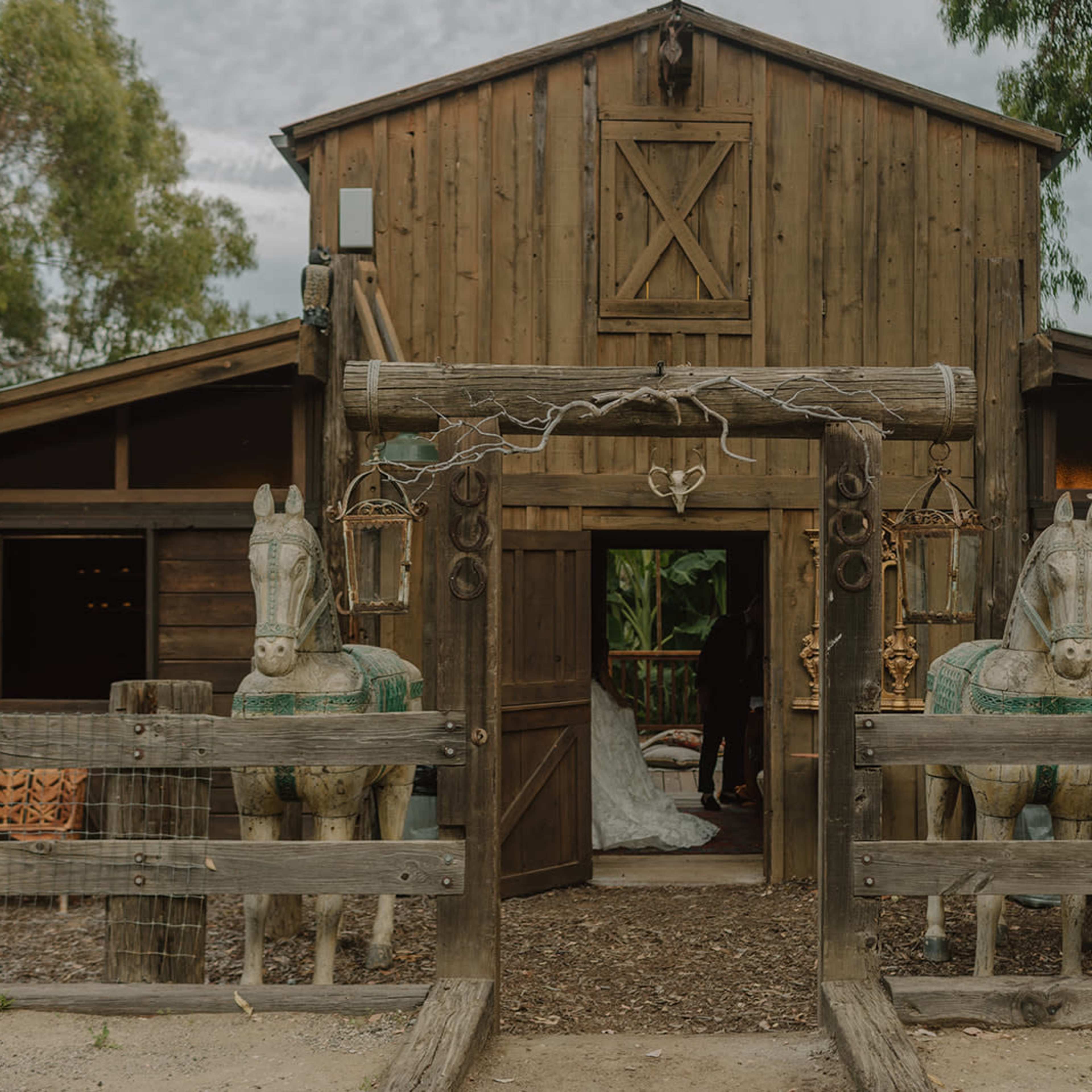 A rustic wooden barn features two carved horse statues at its entrance, flanked by a fenced walkway.