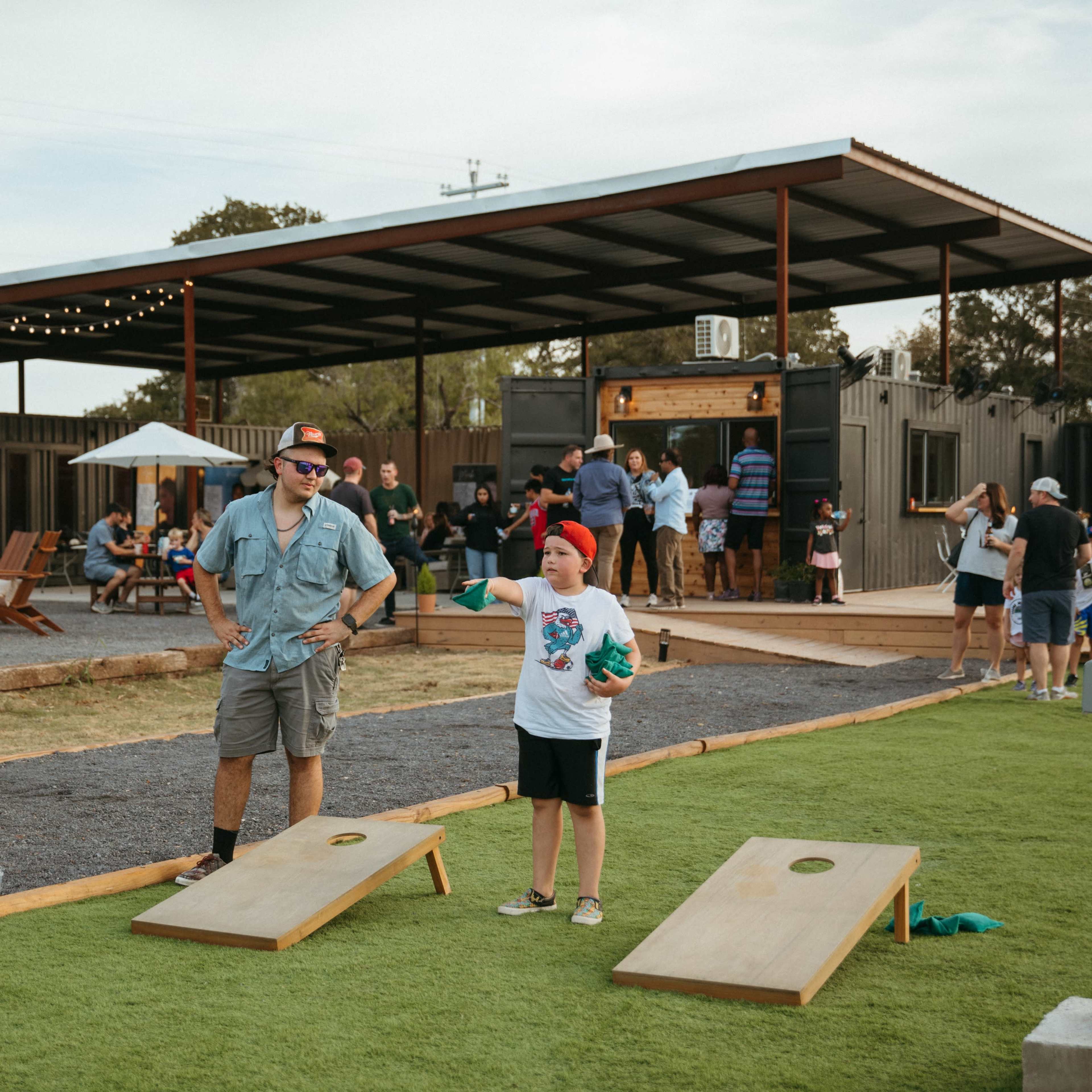 A man and a child play cornhole in a grassy outdoor area while people socialize at a nearby gathering.