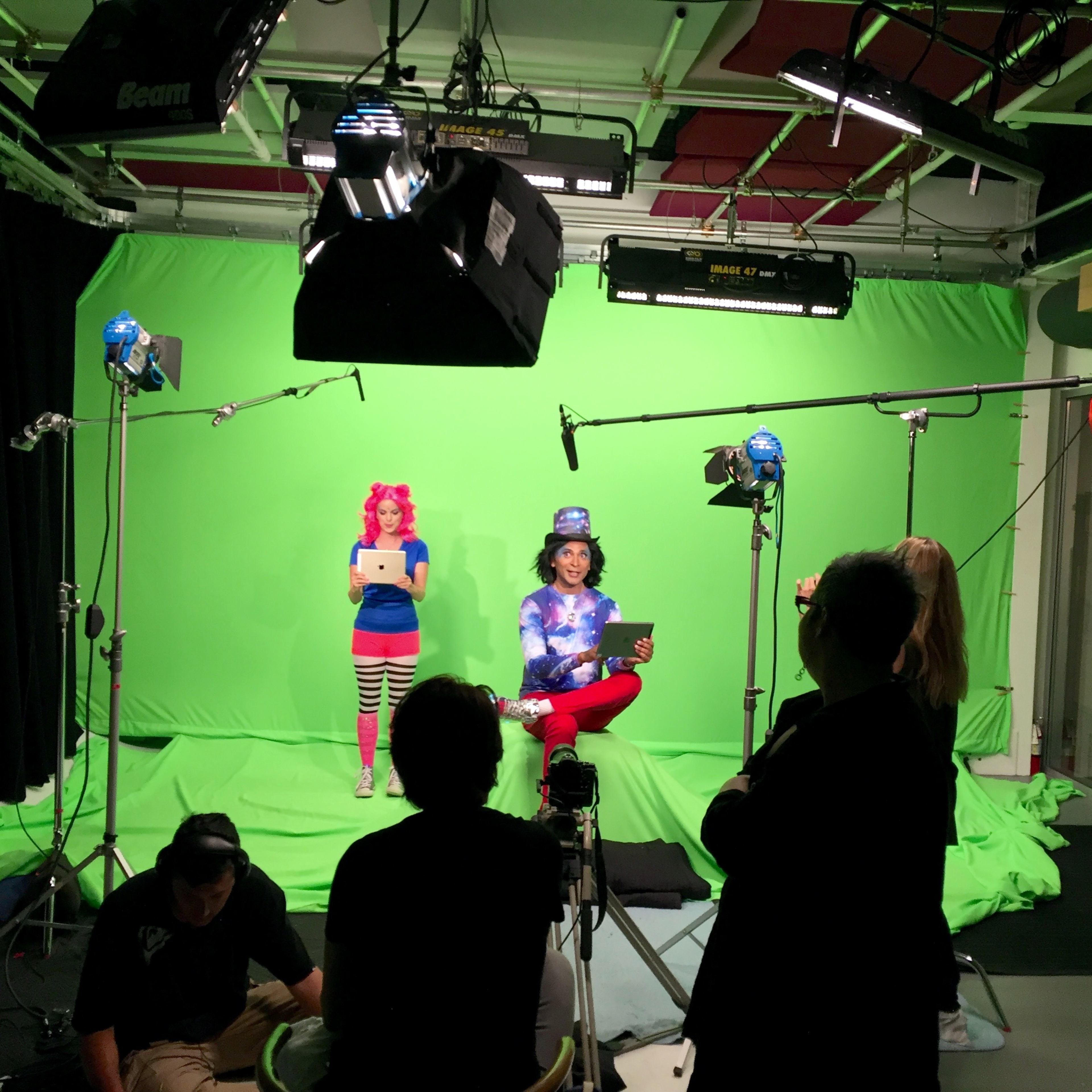 Two performers are seated in front of a green screen, surrounded by cameras and crew members adjusting the lighting and equipment.