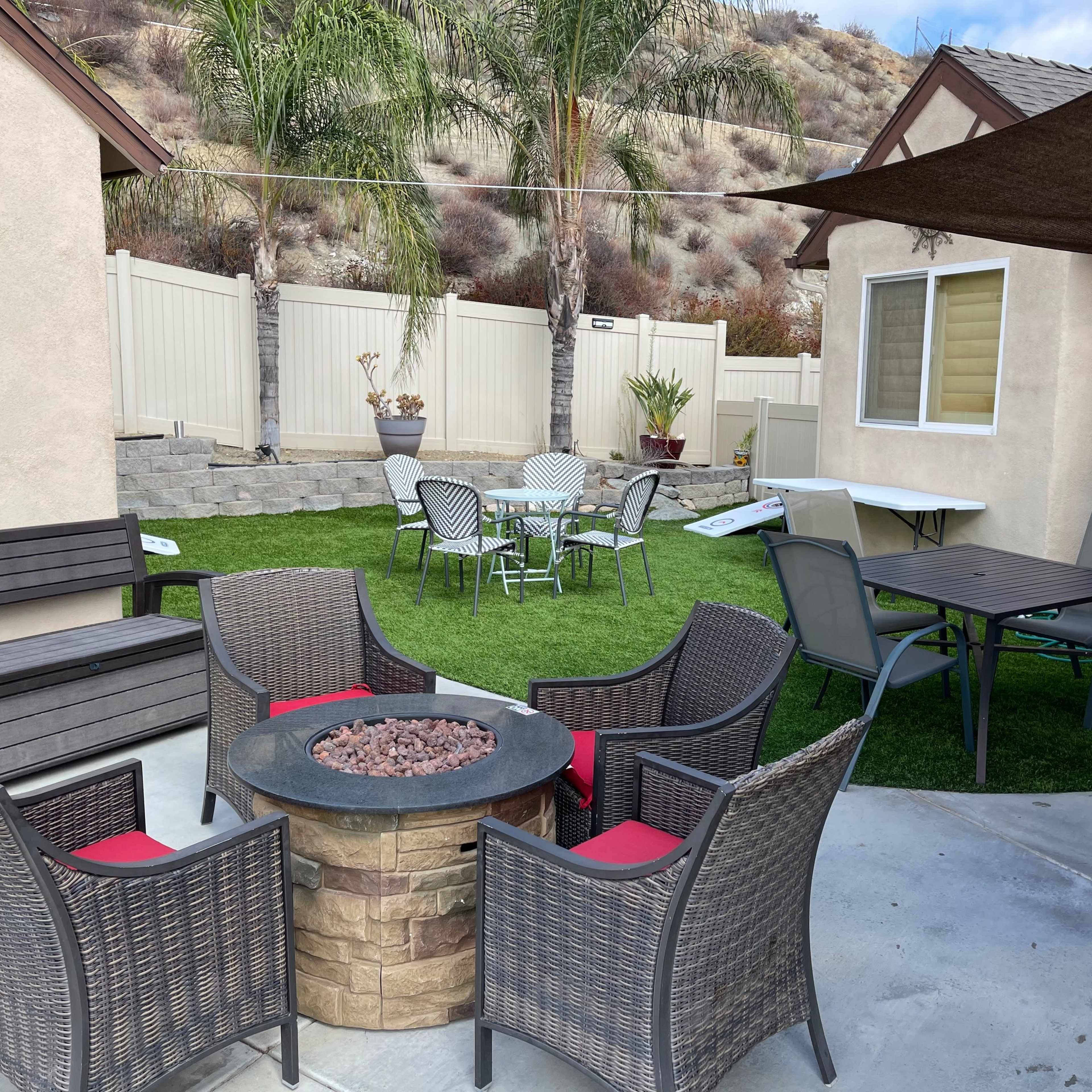 A backyard area featuring a circular fire pit surrounded by four chairs, with additional seating and tables visible on a lawn with a beige wall and palm trees in the background.