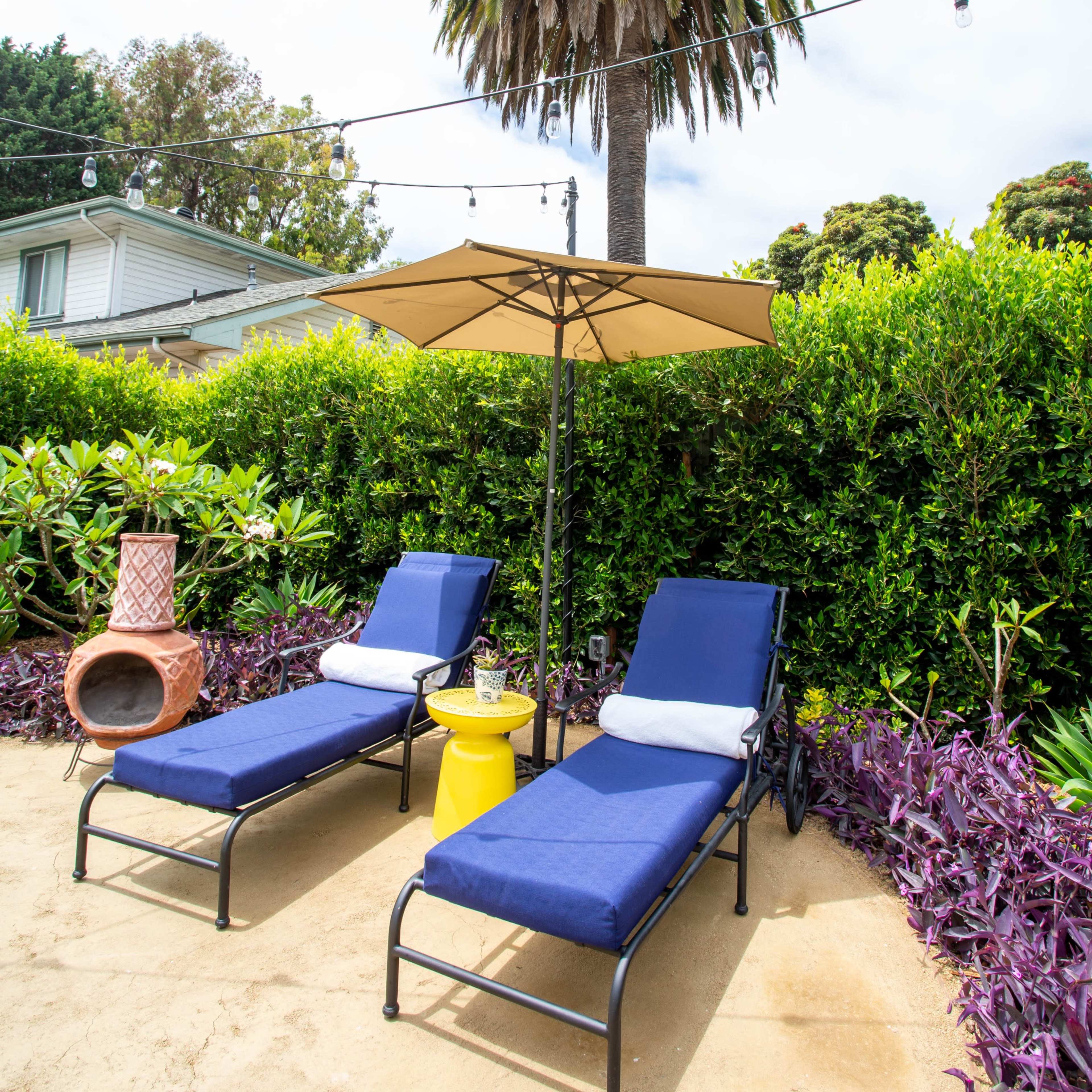 Two blue lounge chairs with an umbrella are placed on sandy ground under a palm tree, surrounded by greenery and purple plants.