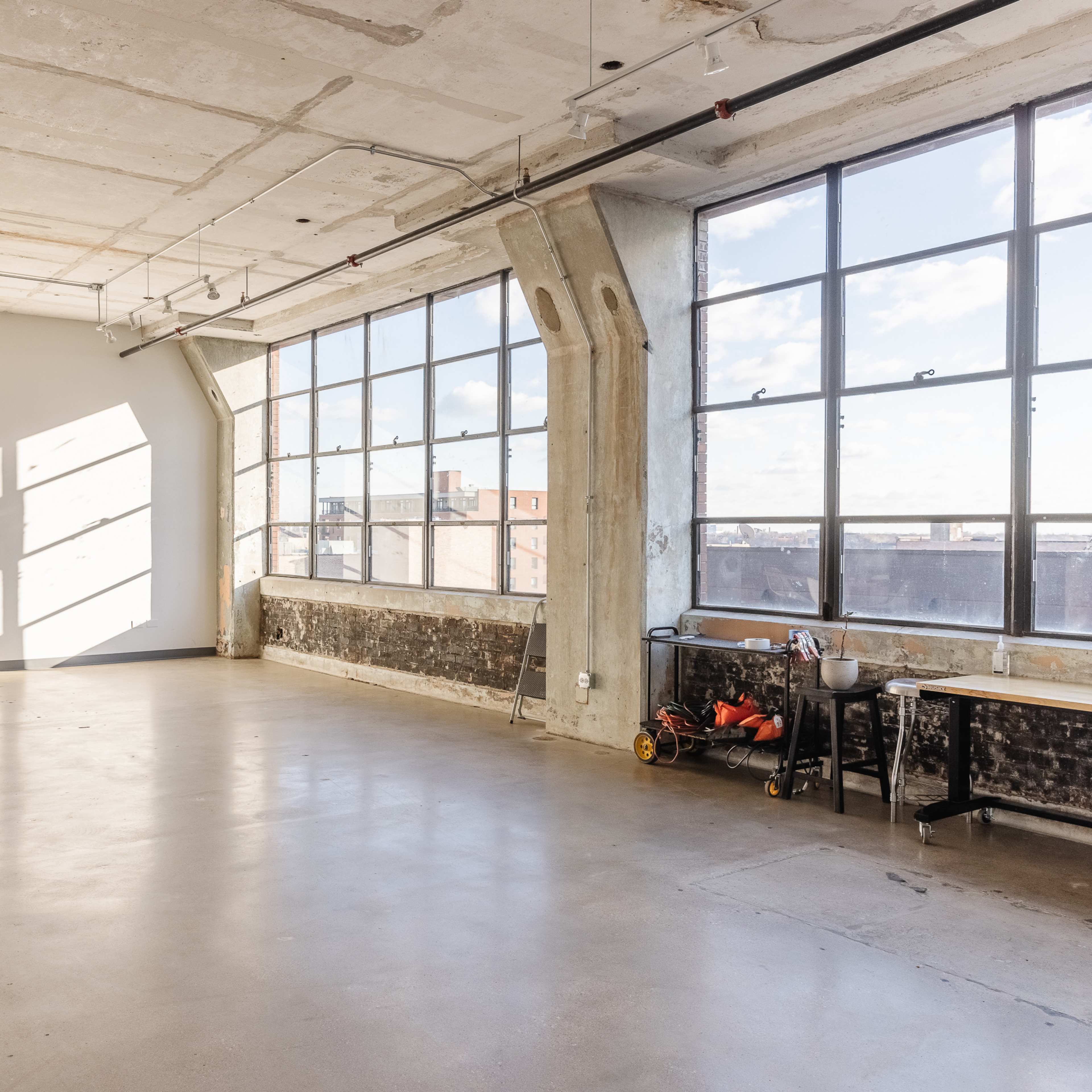 A spacious room with large windows allowing natural light to fill the concrete-floored area, featuring a simple table and a few items along one wall.