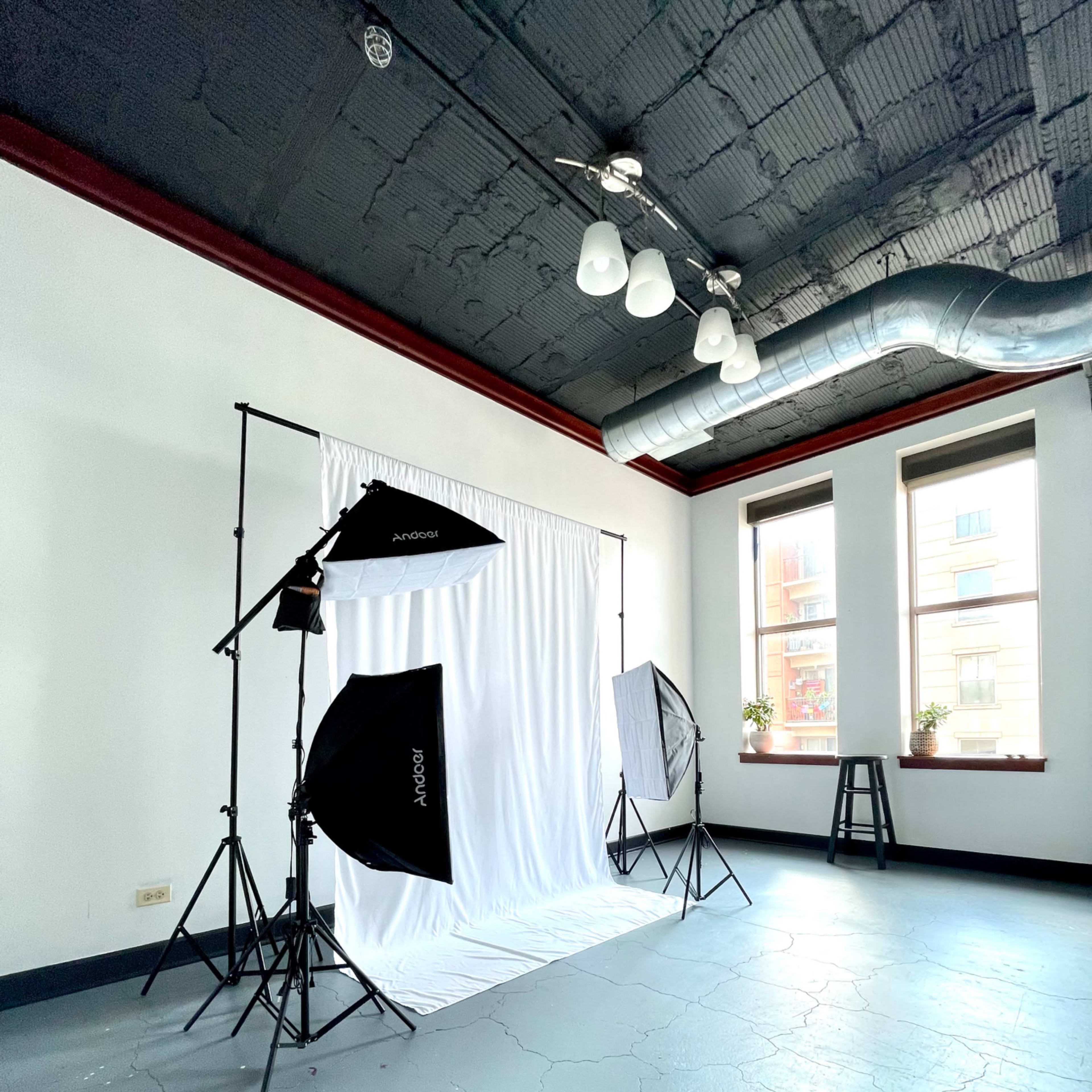 A photography studio with a white backdrop, softbox lights on stands, and a wooden stool in a brightly lit room.