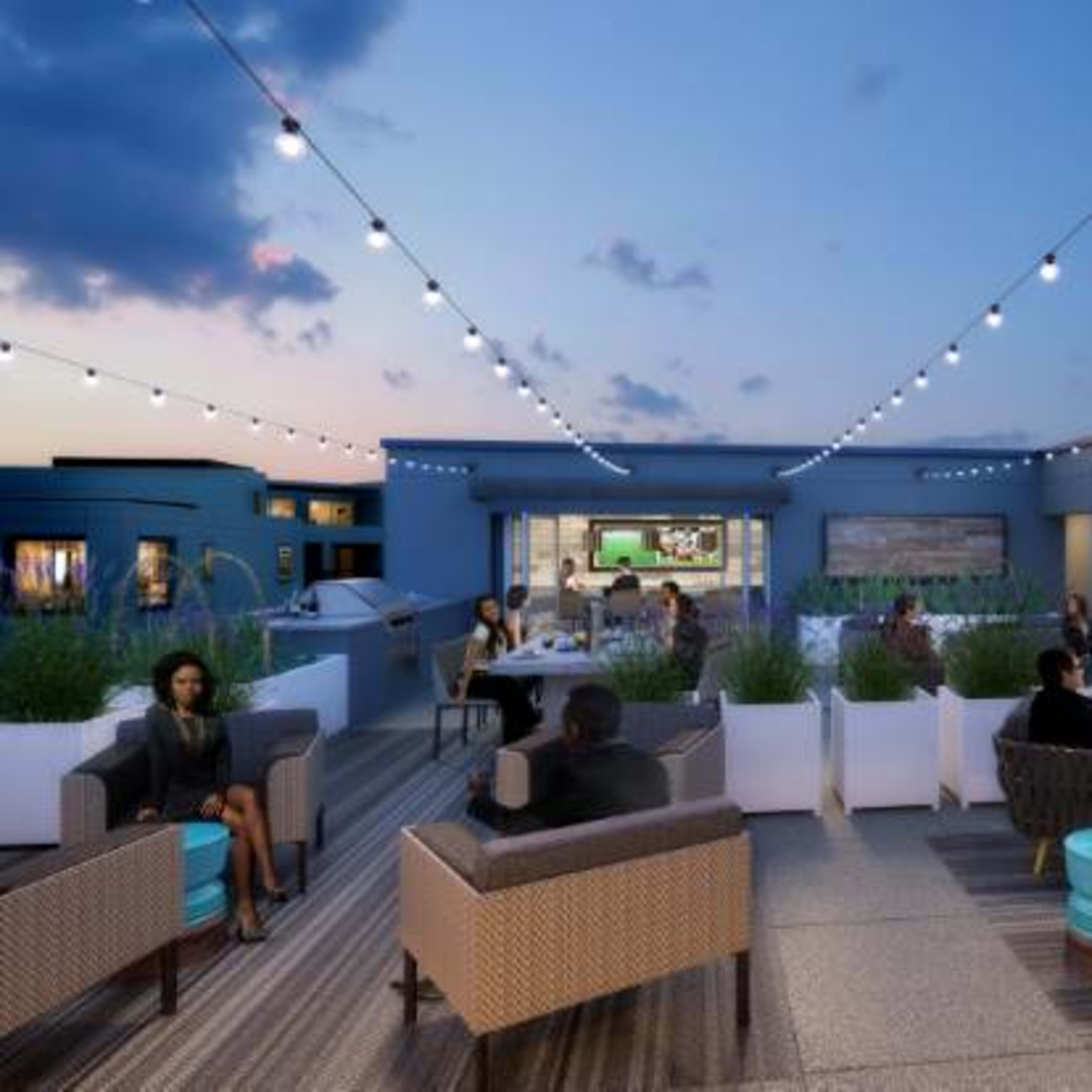 A rooftop lounge area features comfortable seating, string lights, and a modern fireplace, with people socializing in the evening.