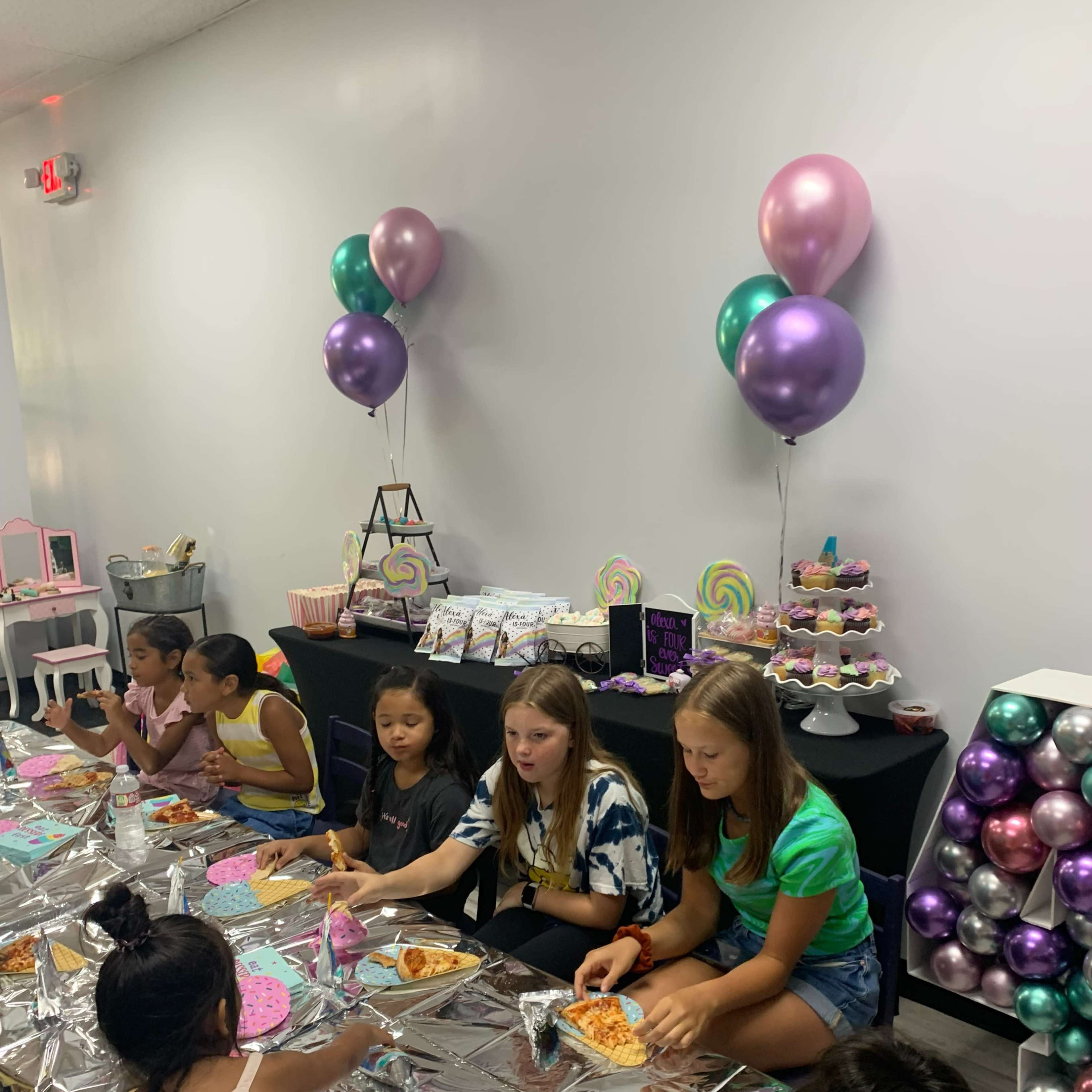 A group of young girls sits at a decorated table, enjoying pizza and snacks at a colorful birthday party.