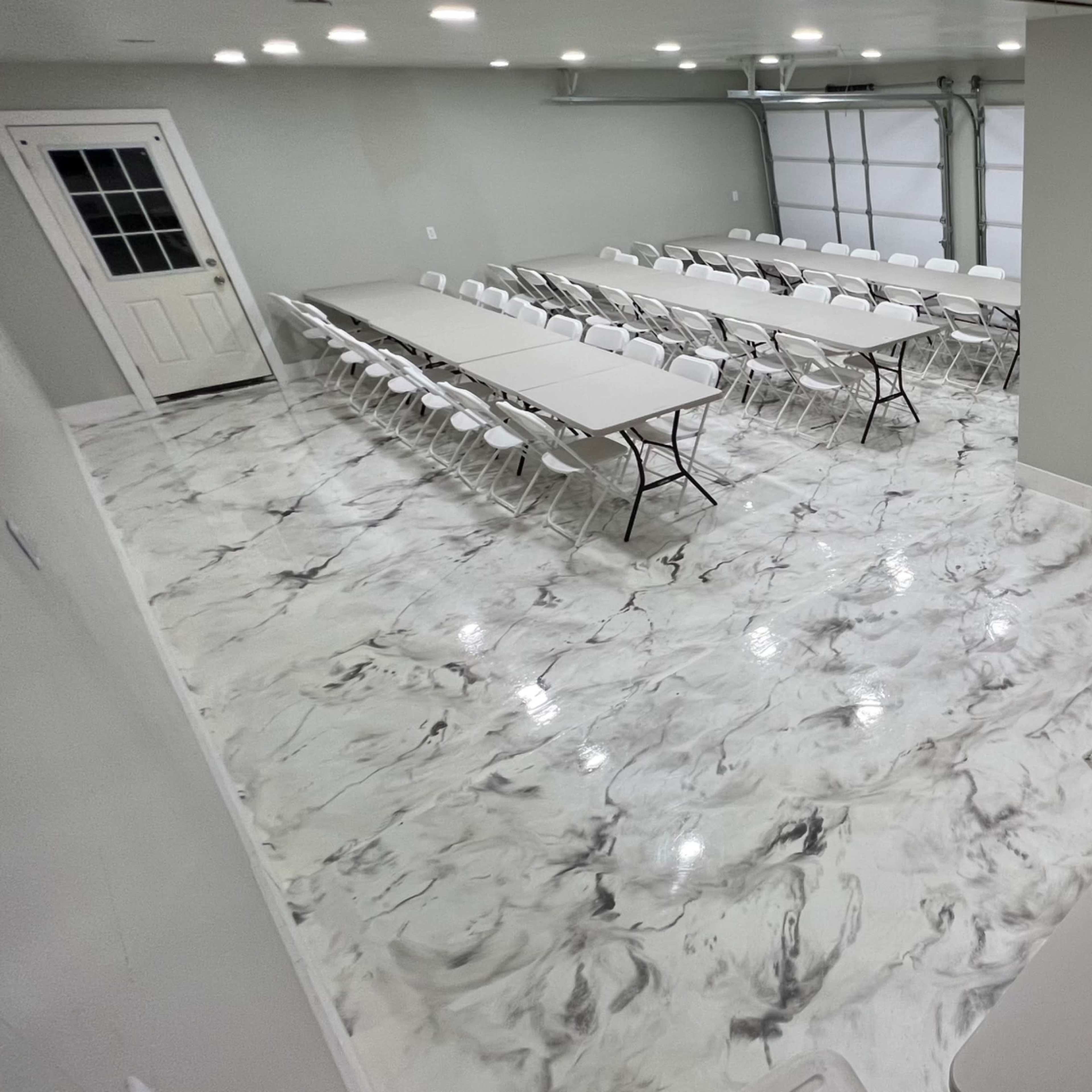 A large room set up for an event, featuring multiple tables arranged in rows, white chairs, and a decorative balloon arrangement in the corner.