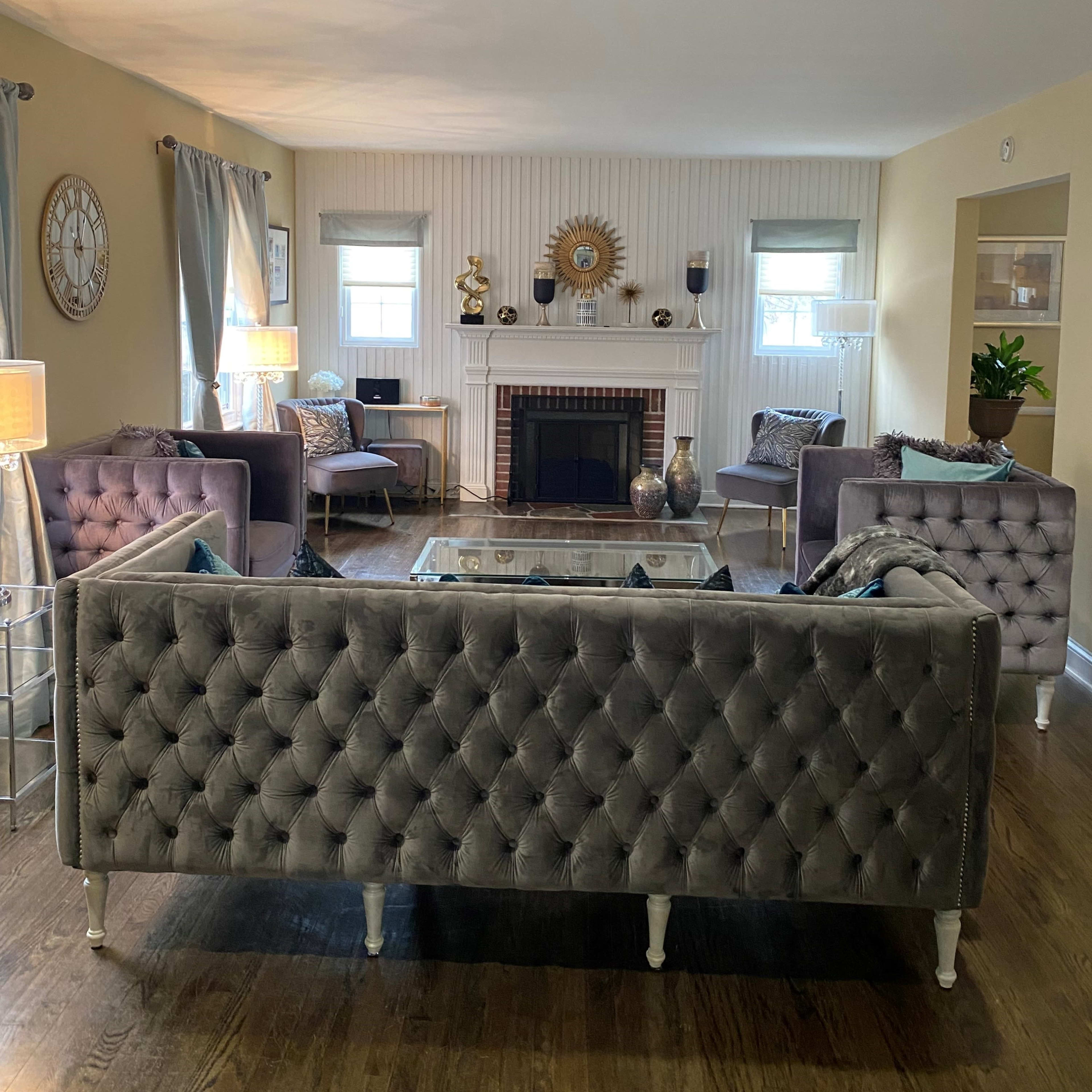 A living room features tufted gray sofas, a glass coffee table, a decorative fireplace, and large windows with light-colored curtains.