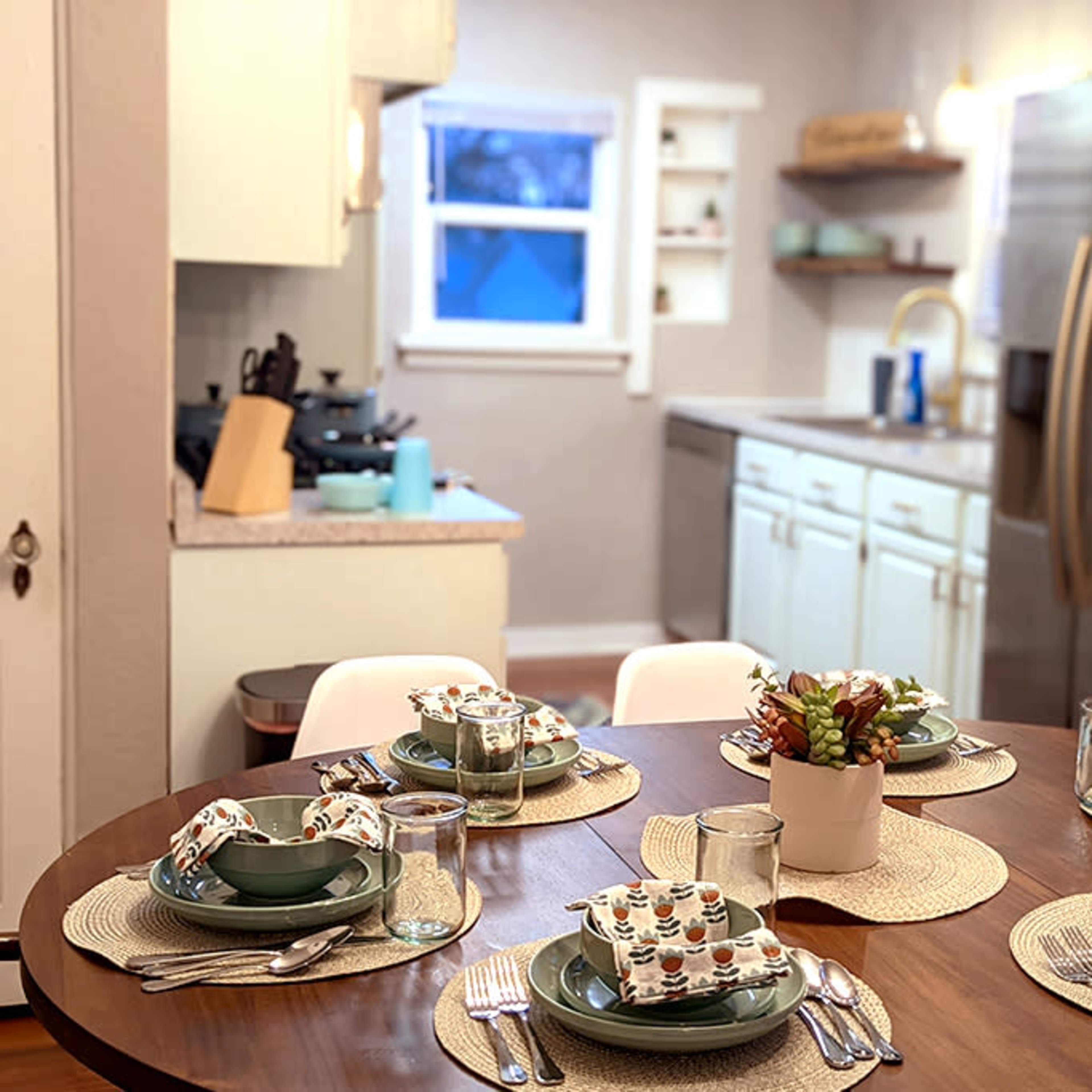 A dining table is set with plates and glasses, accompanied by a kitchen visible in the background.