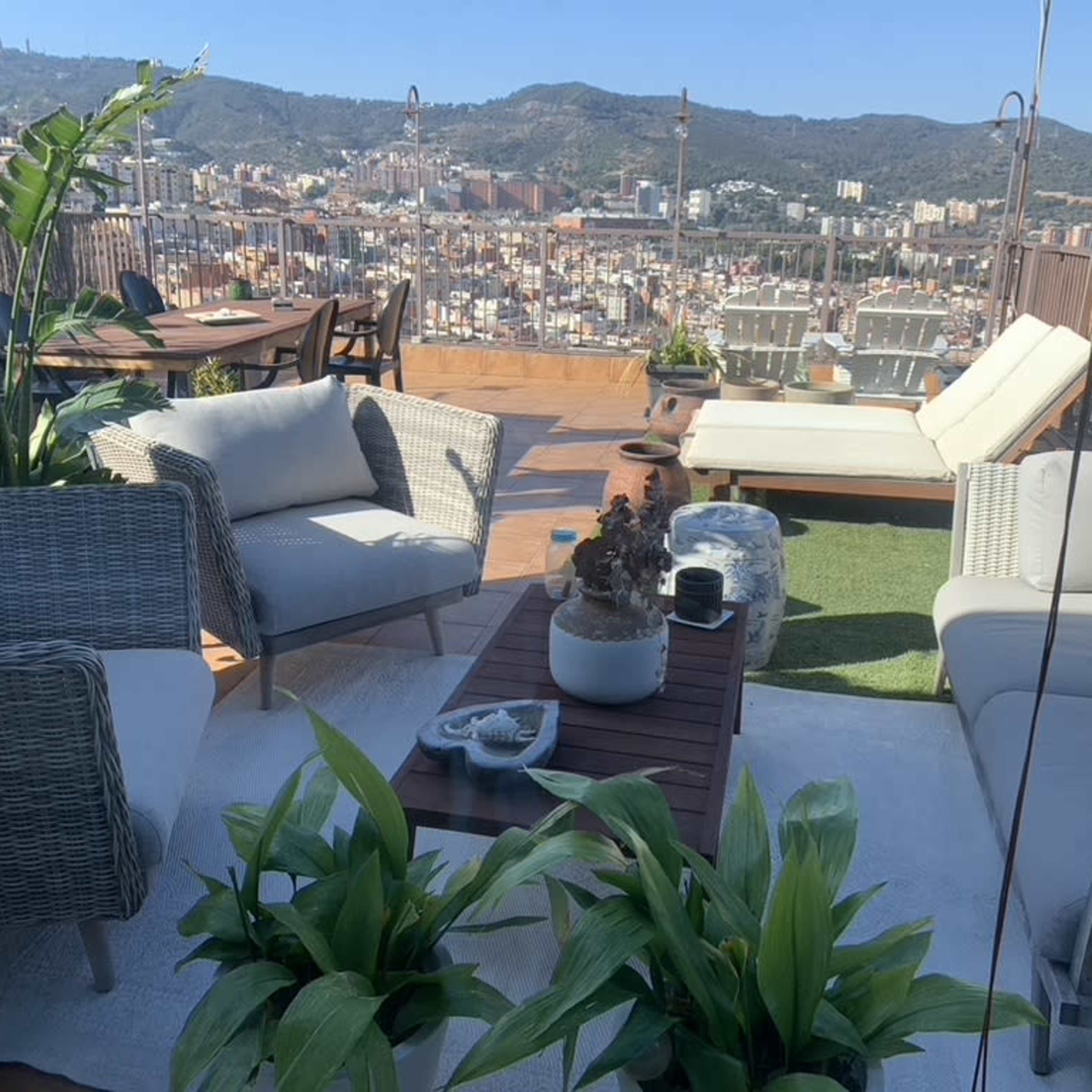 A rooftop terrace with seating areas, potted plants, and a view of a city landscape in the background.