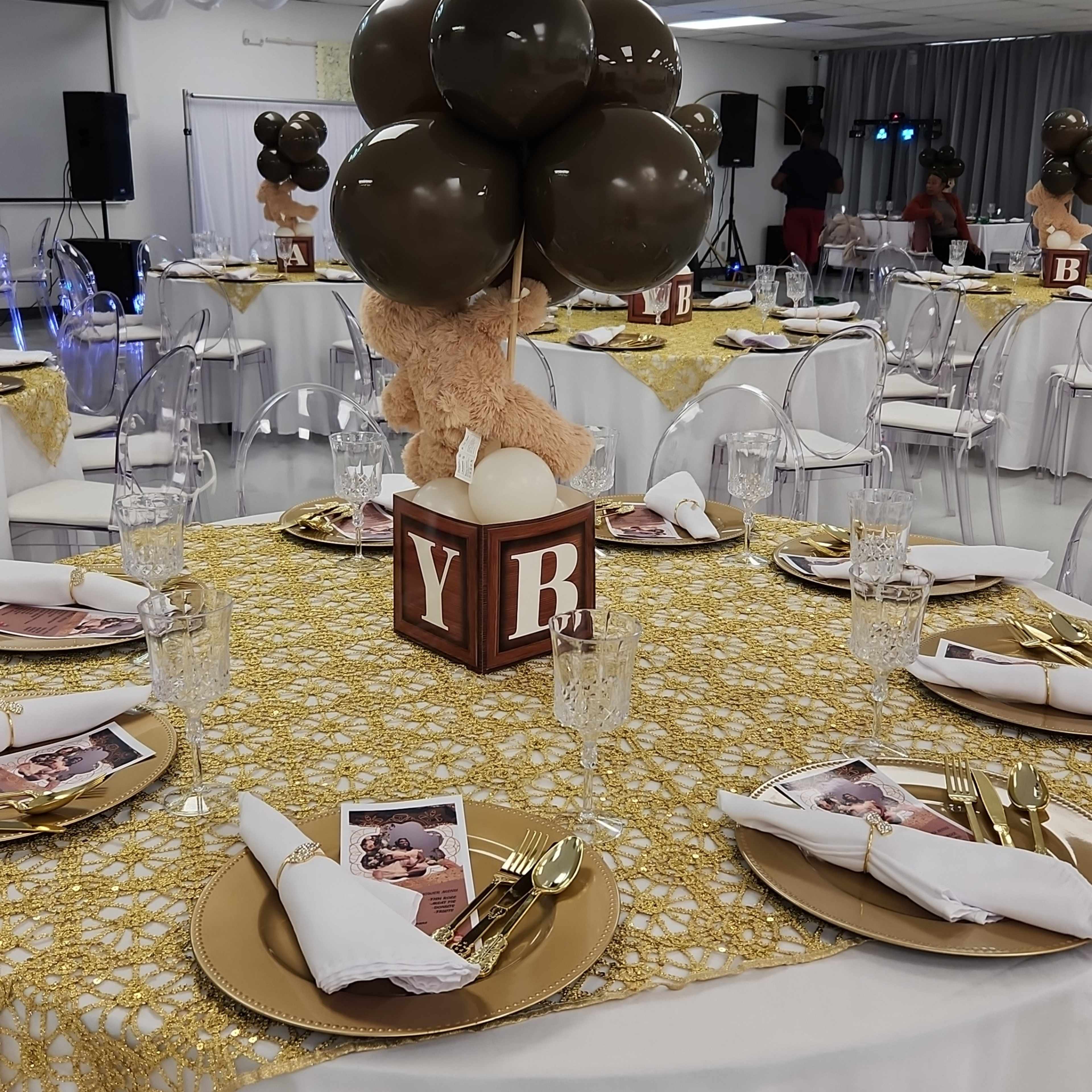 A decorative table is set for a celebration, featuring a gold tablecloth, clear chairs, and a centerpiece with balloons and a teddy bear.