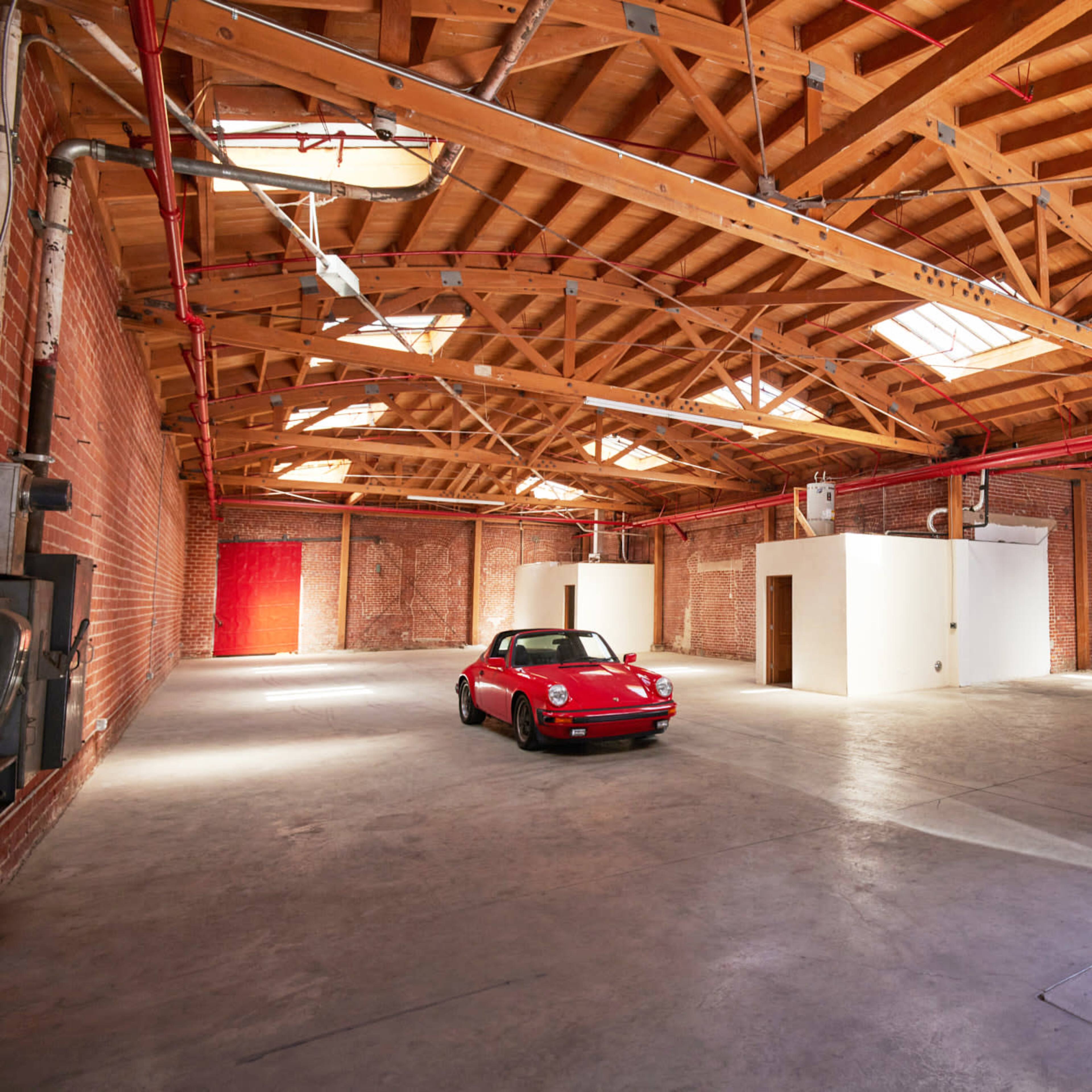 A red sports car is parked in a spacious warehouse with wooden beams and white walls.