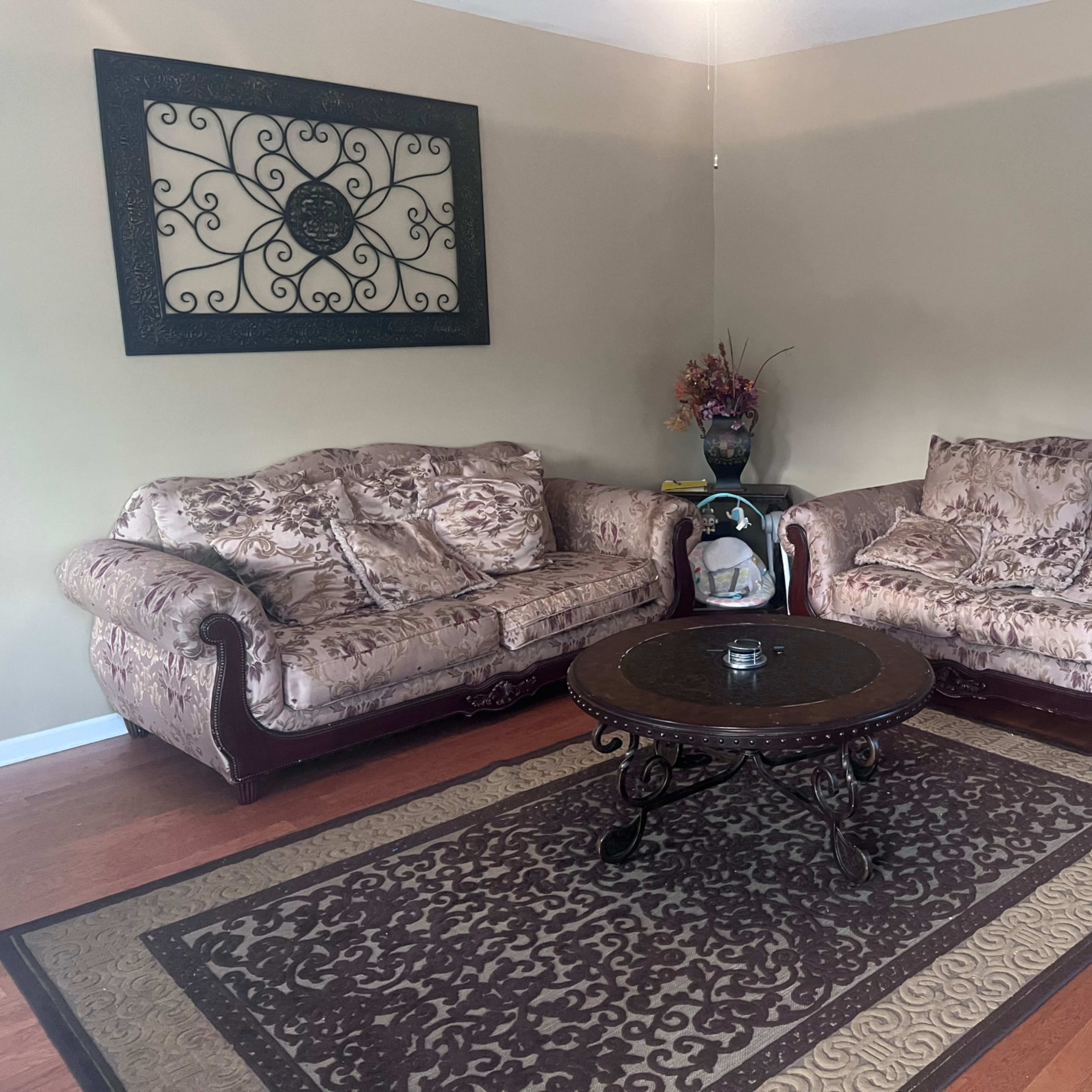 A living room with two ornate sofas facing each other, a round coffee table in the center, and a decorative wall hanging above.