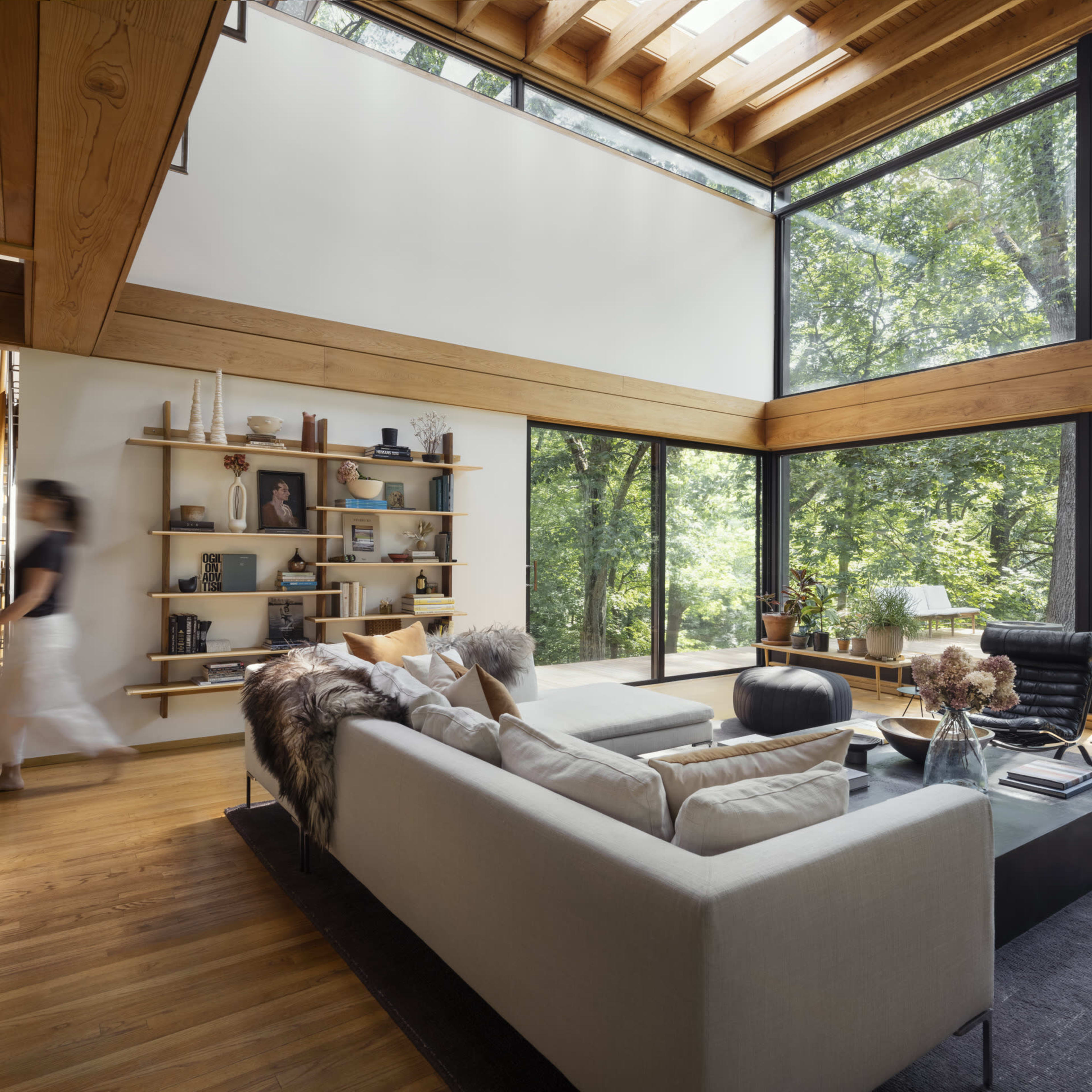 A modern living room features large windows with a view of trees, a sectional sofa, and minimalist shelving.