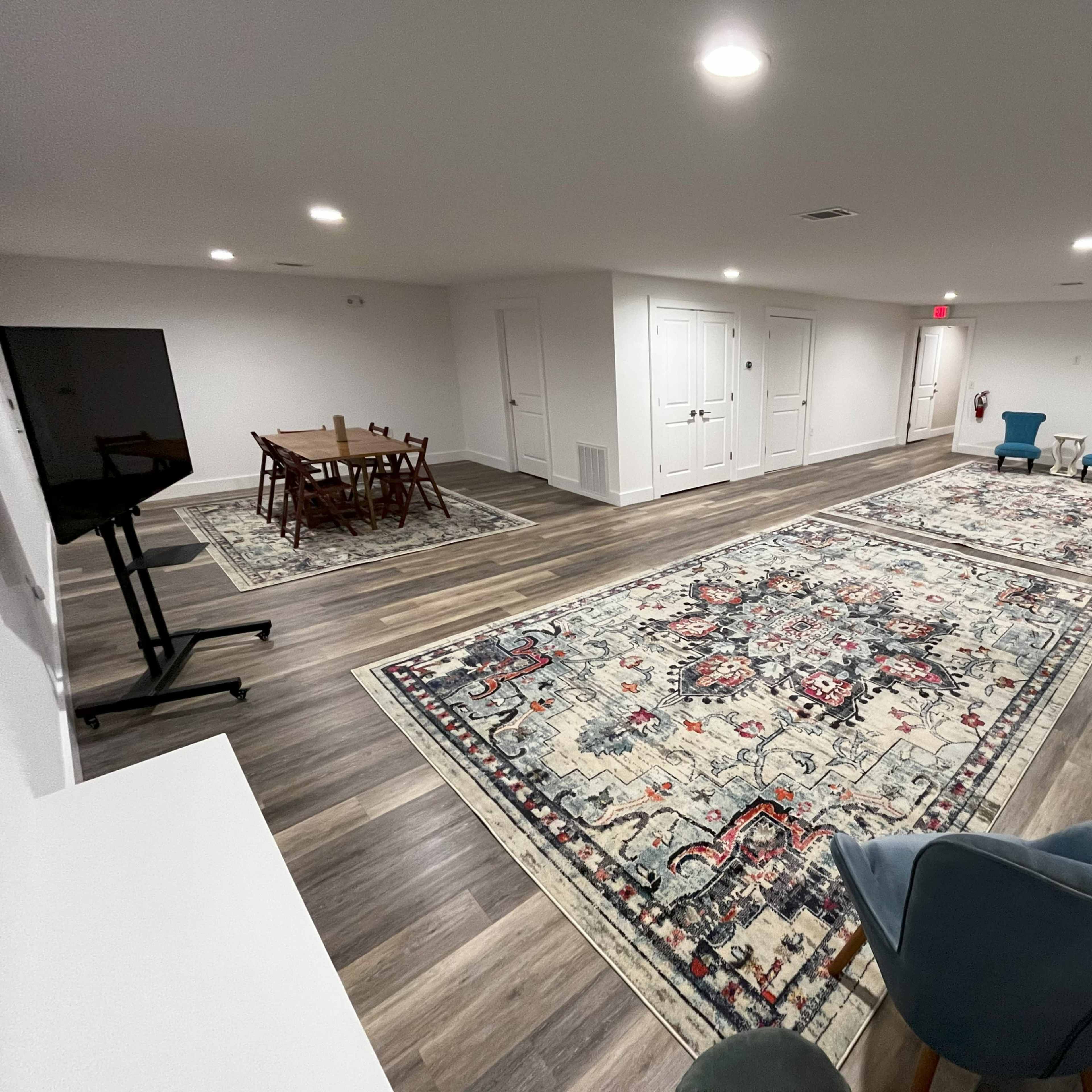 A spacious room with hardwood flooring, two large area rugs, a dining table with chairs, a television on a stand, and two blue accent chairs.