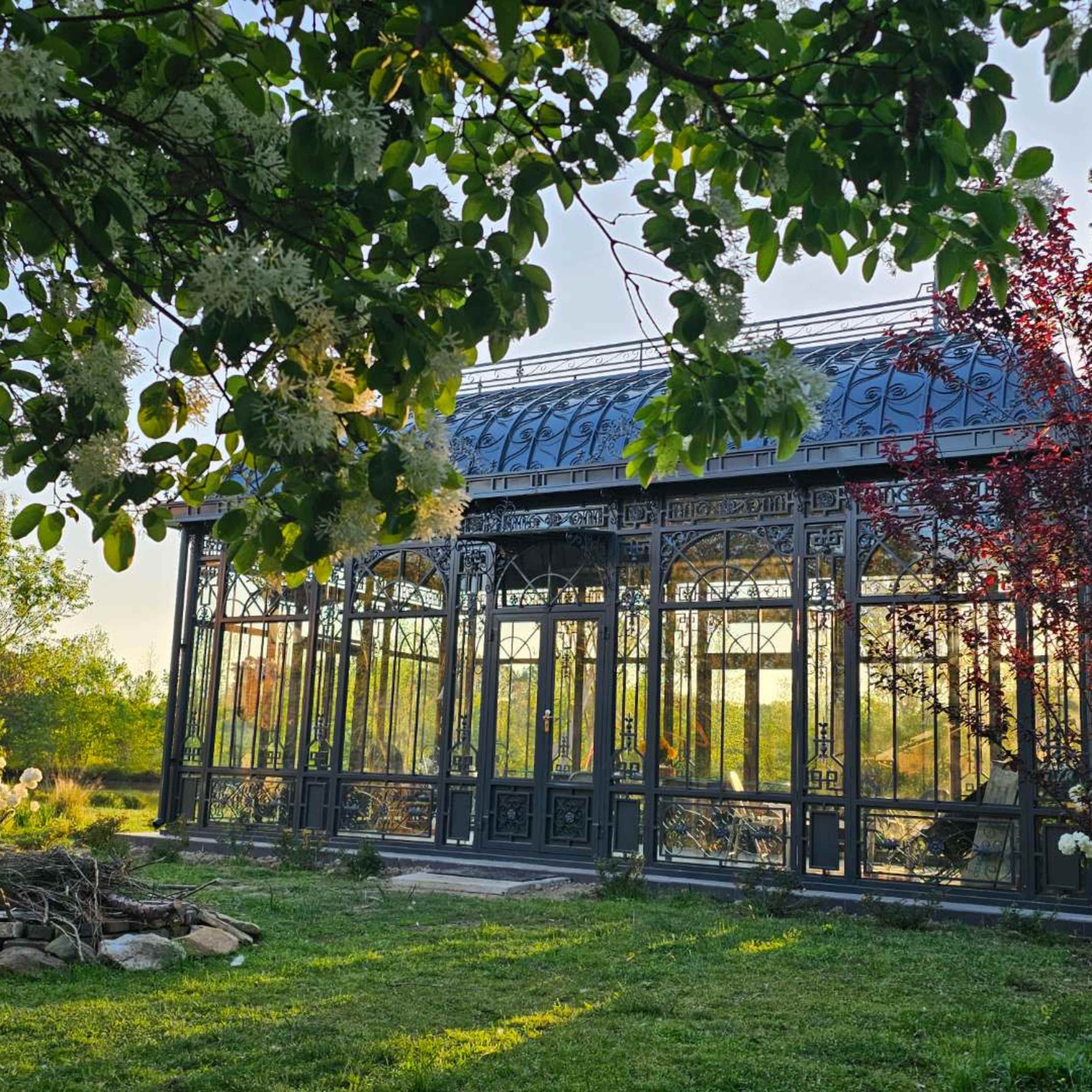 A glass conservatory with intricate metalwork is situated in a grassy area, surrounded by trees and plants.