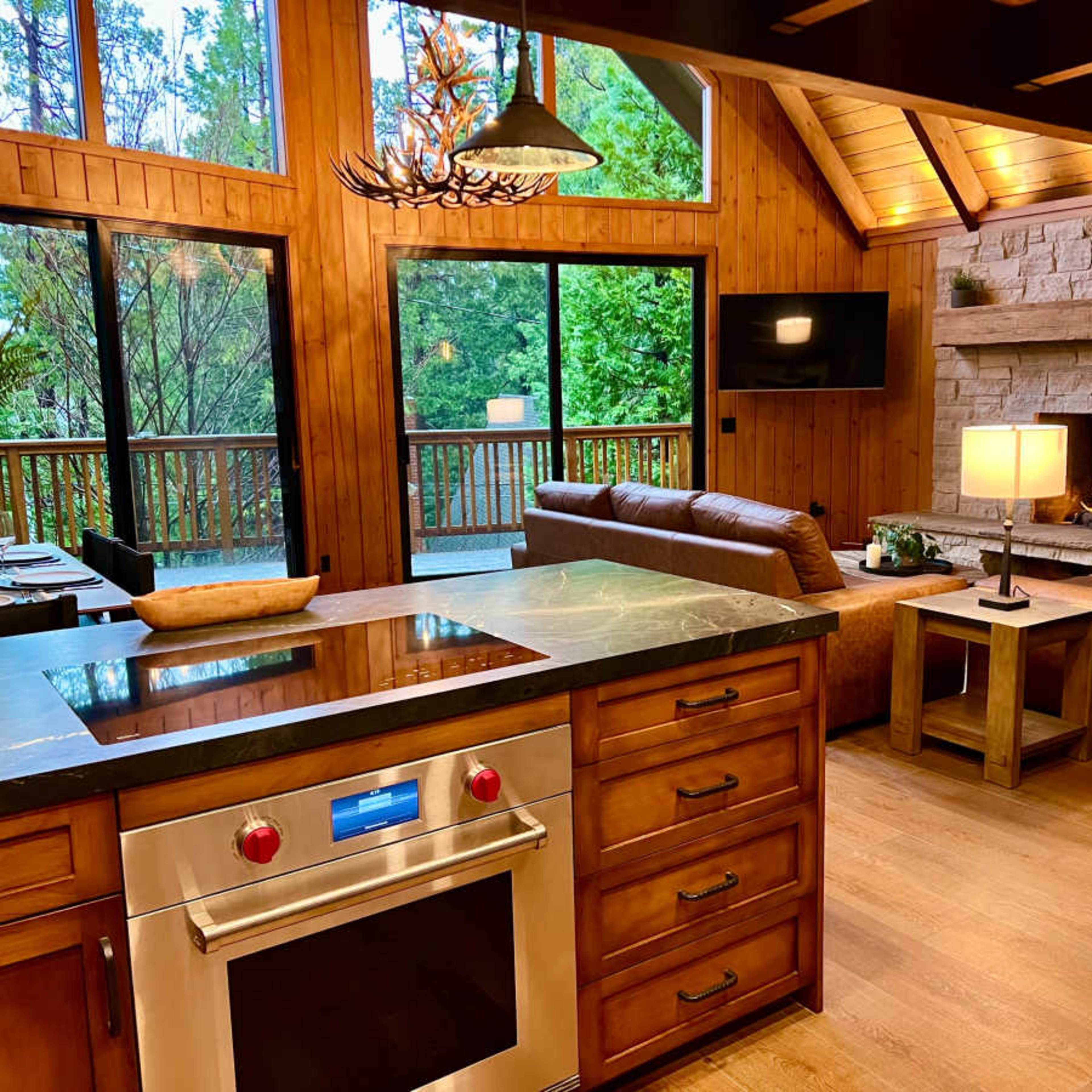 A modern kitchen with a stainless steel oven opens into a cozy living area featuring a stone fireplace and large windows overlooking a wooded view.