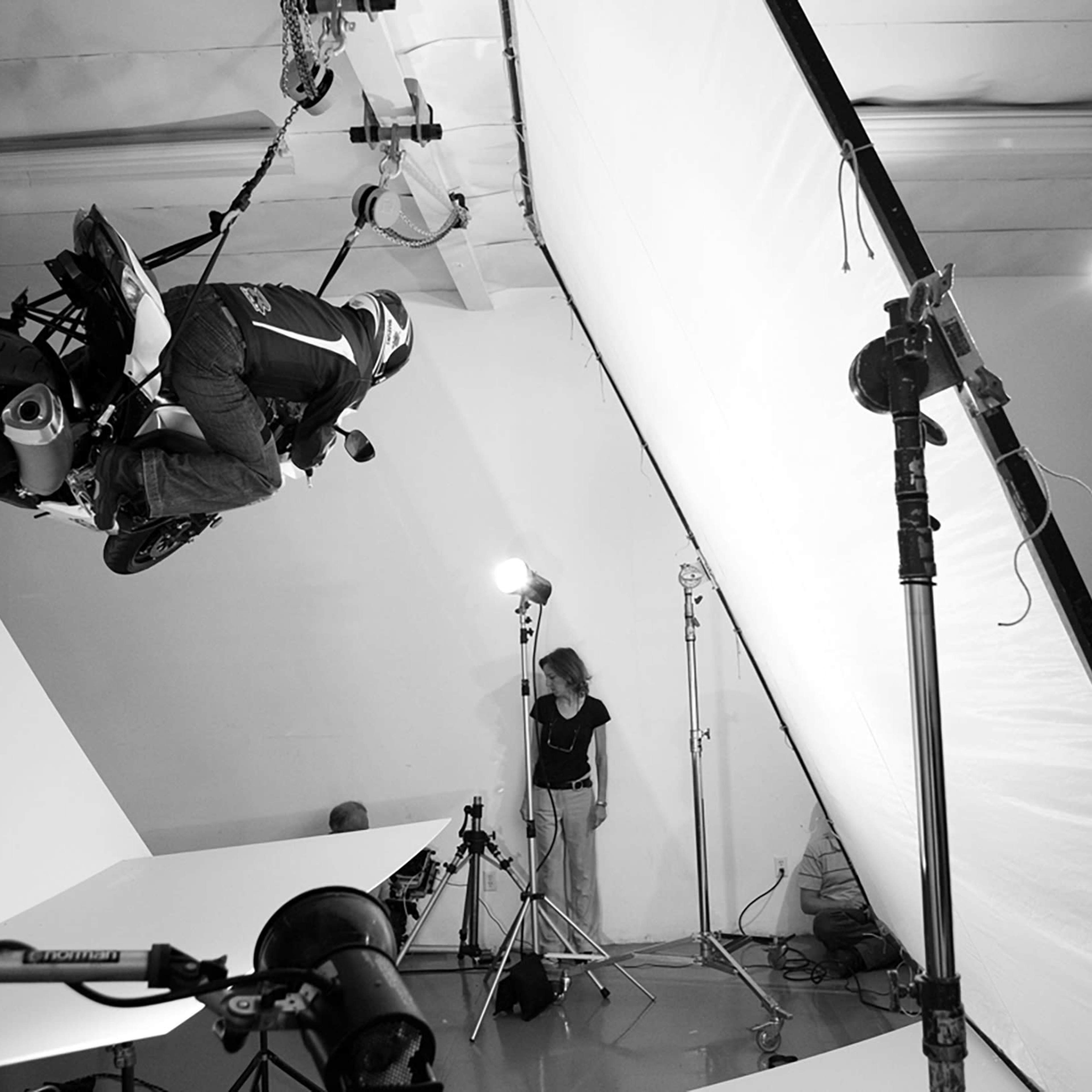 A motorcycle rider performs a stunt in a photography studio, surrounded by lighting equipment and backdrops.