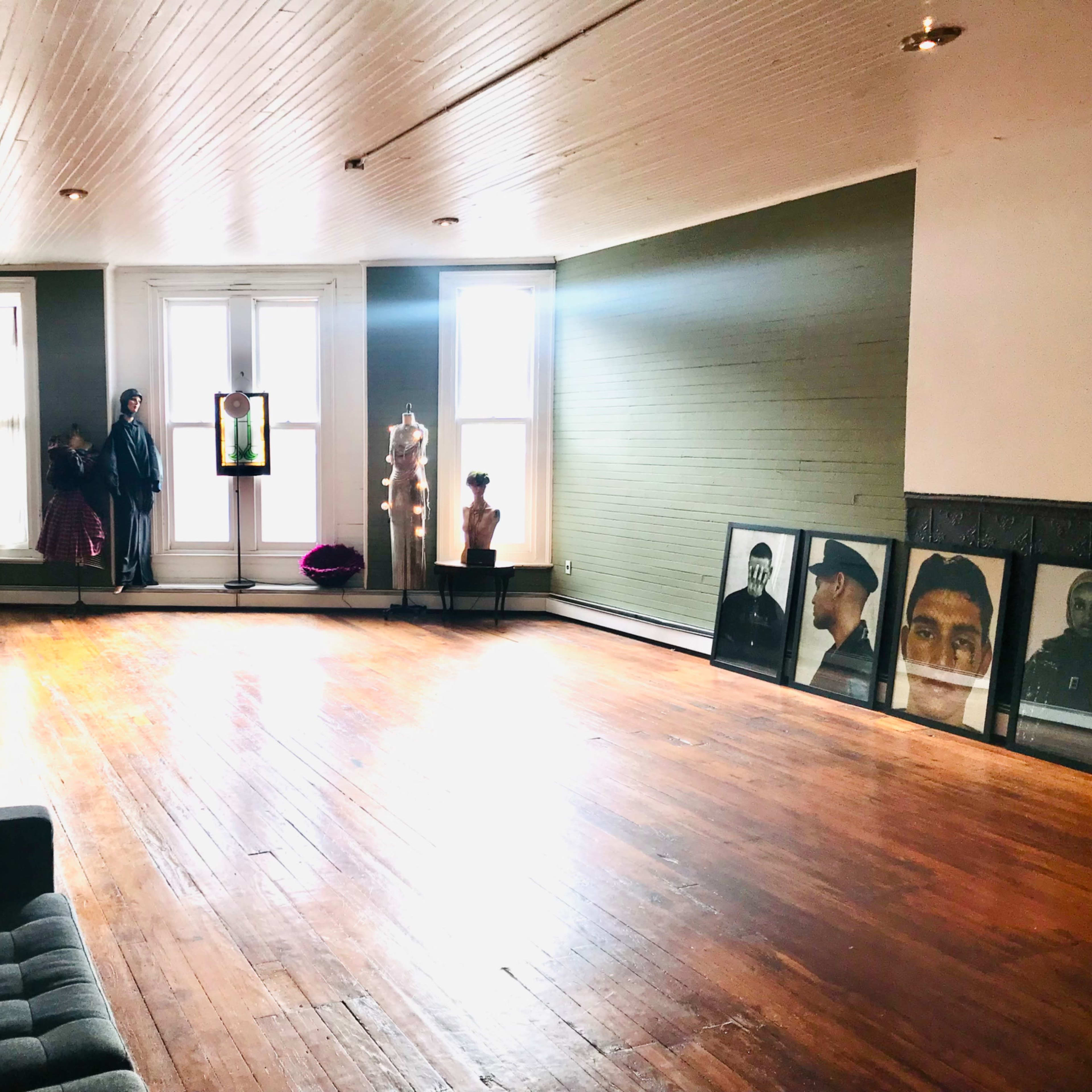 A spacious, empty room with wooden floors, large windows, and various artworks and mannequins displayed along the walls.
