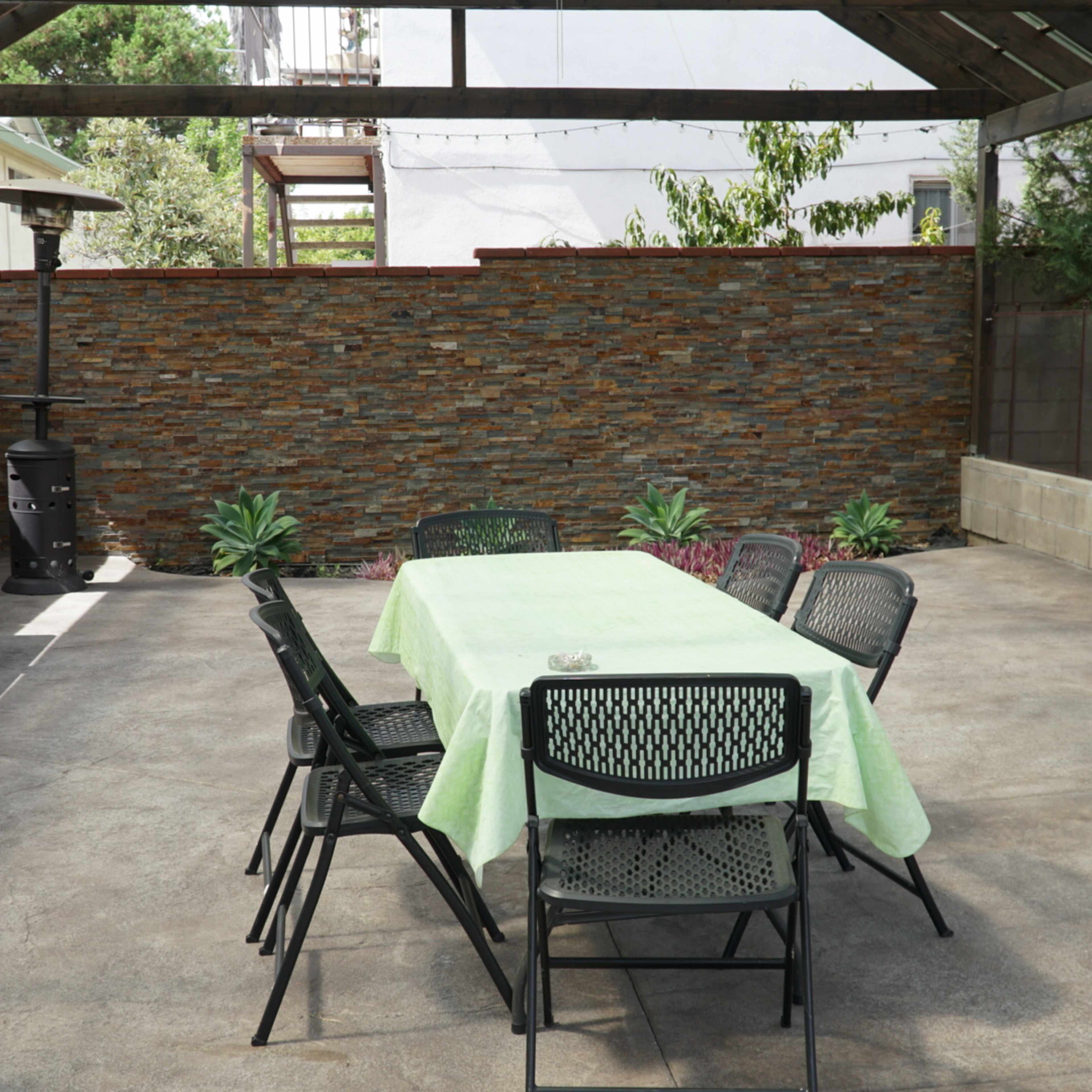 An outdoor patio features a table covered with a green tablecloth surrounded by black chairs, with a stone wall in the background.