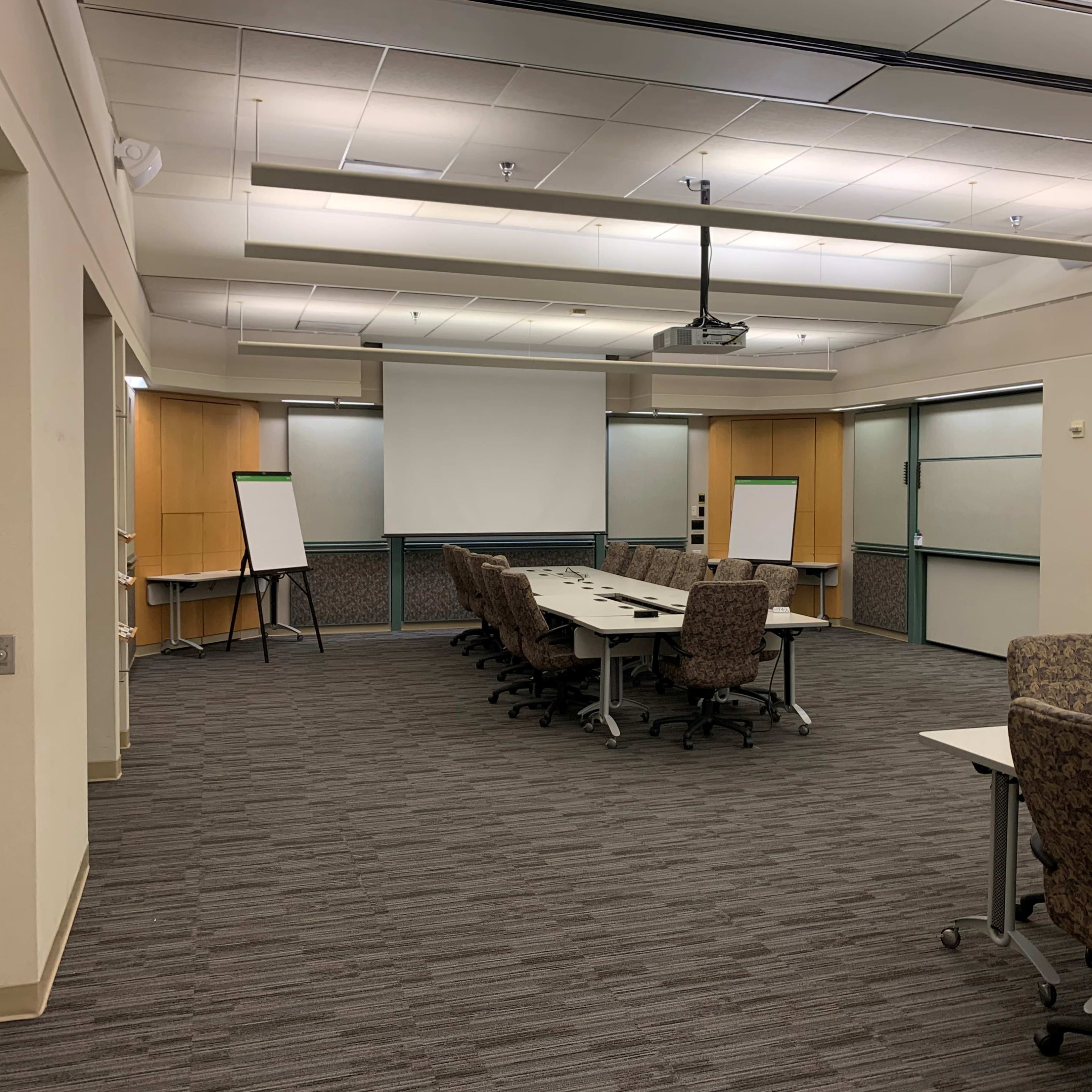 A conference room is set up with a large table surrounded by rolling chairs, audiovisual equipment, and whiteboards along the walls.