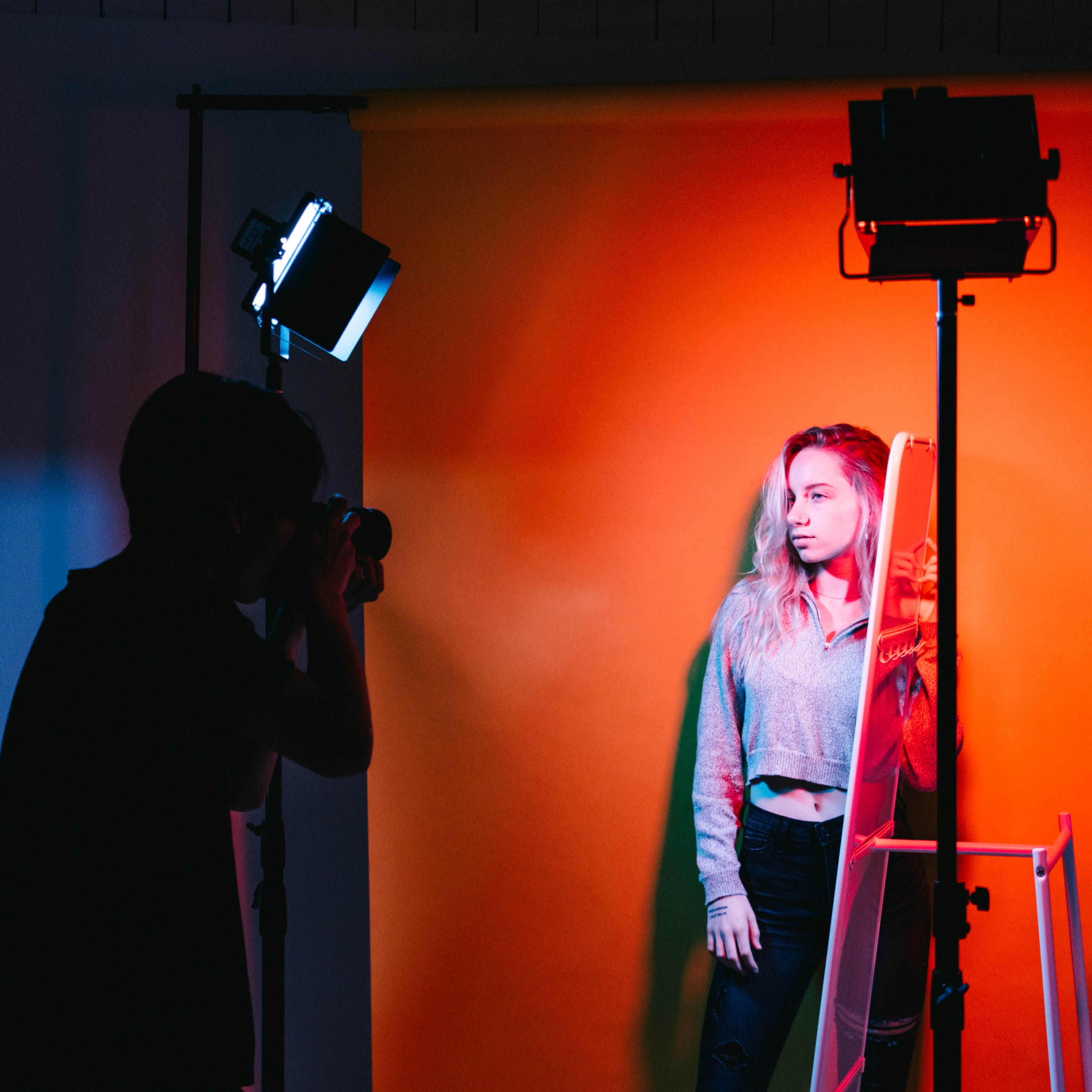A photographer takes a picture of a model standing against a brightly colored background with studio lights illuminating the scene.