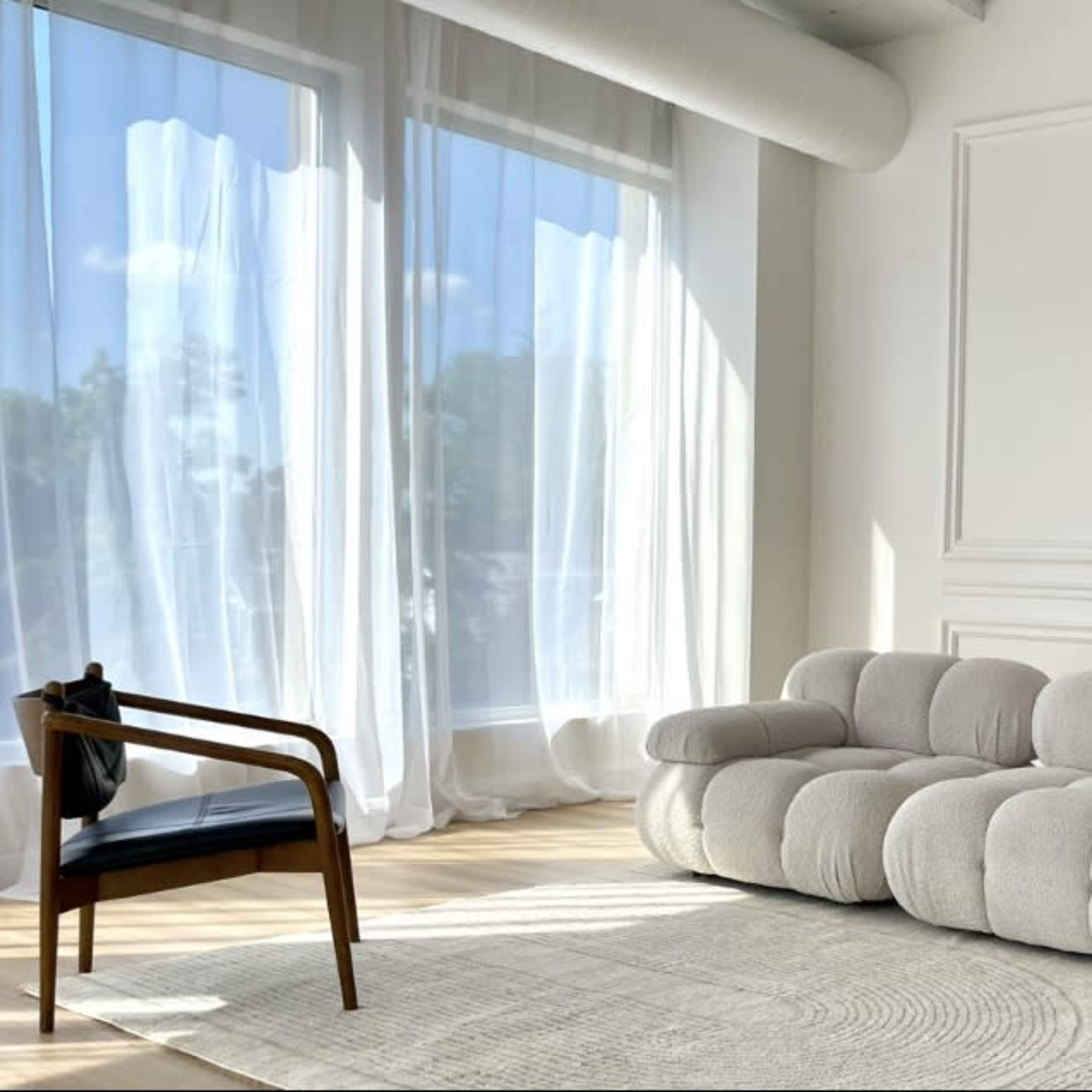 A modern living room with a large, curved beige sofa, two wooden chairs, and large windows covered with sheer white curtains.