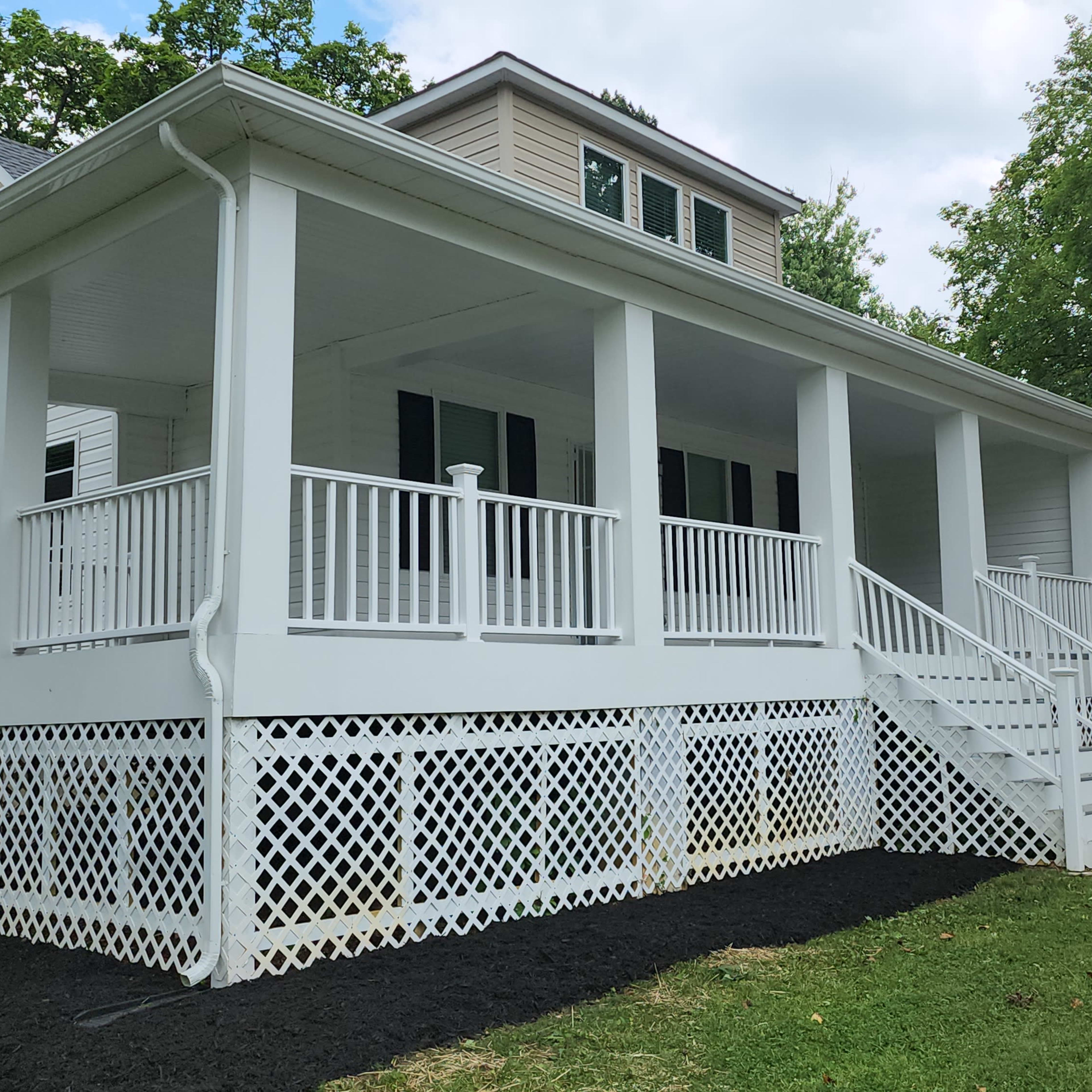 A white house with a spacious front porch, featuring a lattice skirting and surrounded by green grass and trees.