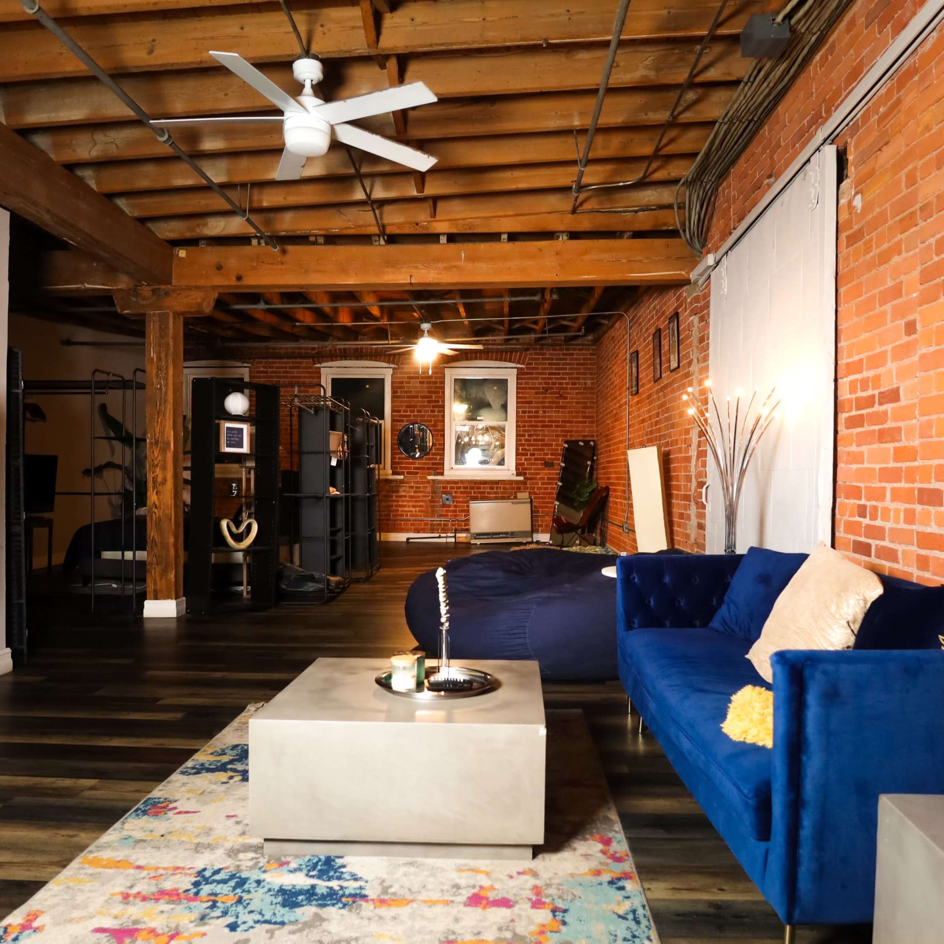 A spacious, modern living area with exposed brick walls, hardwood flooring, a large television on a stand, and a blue couch with yellow accents.