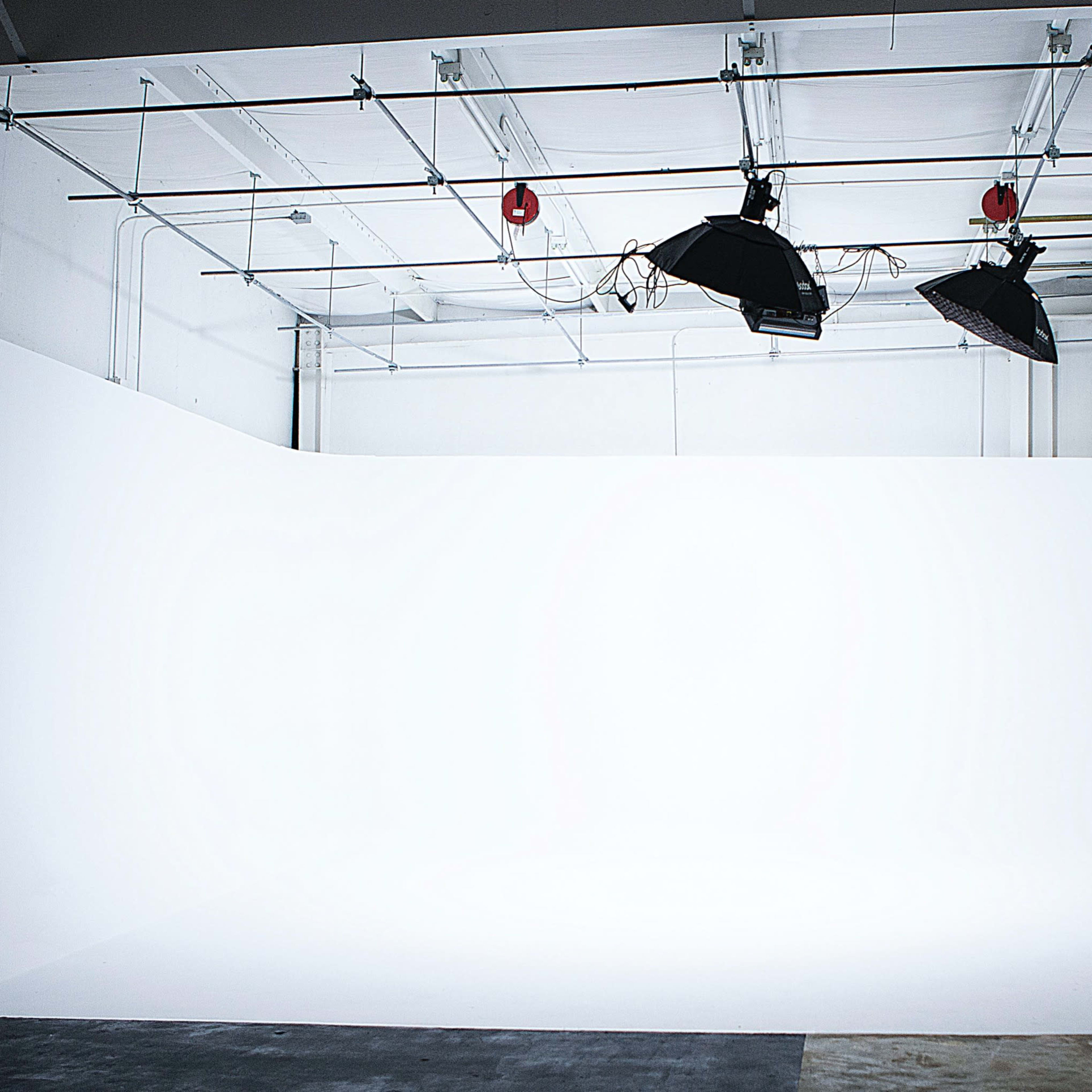 A photography studio with a large white backdrop, two overhead lights, and a softbox on a stand.