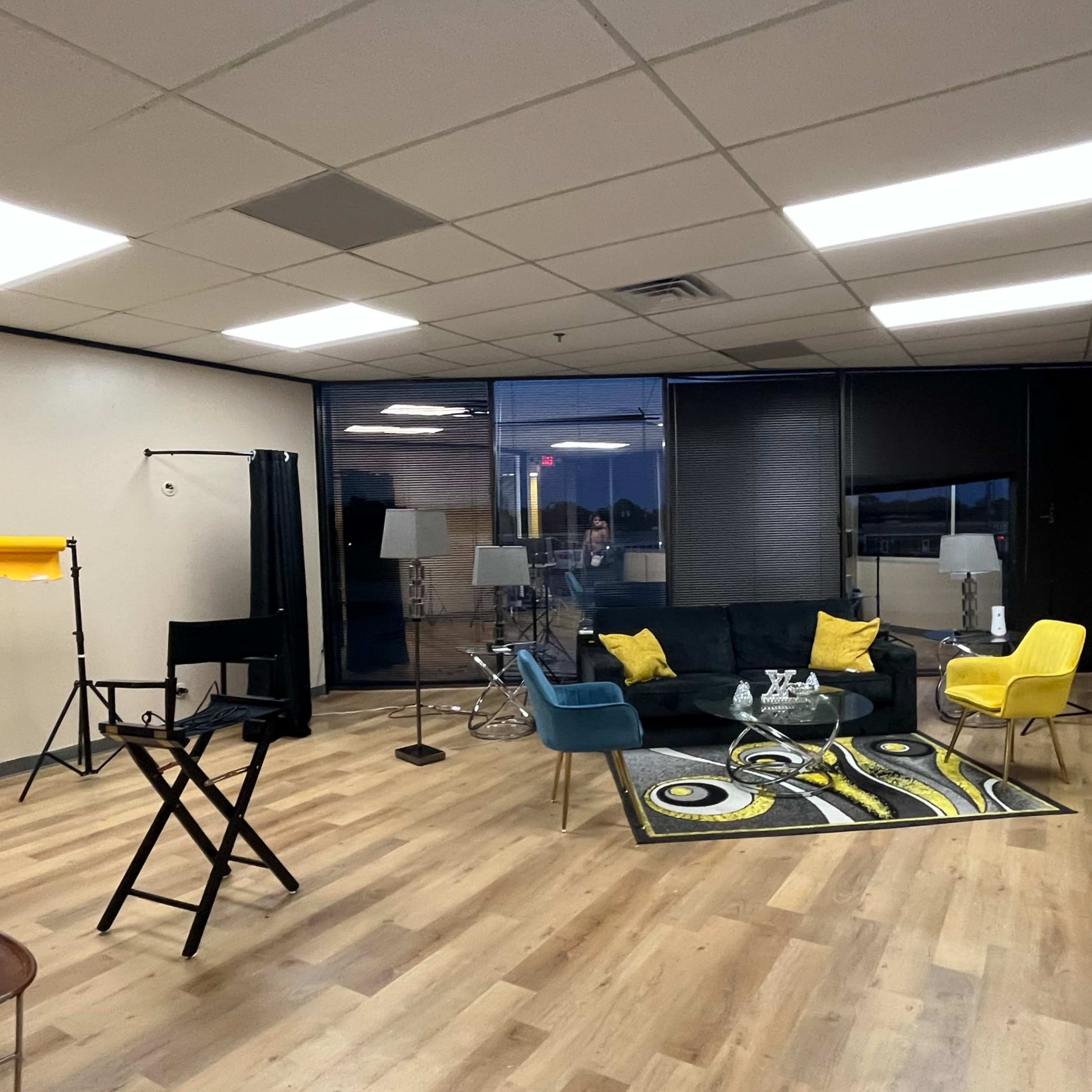 A modern studio space with a black sofa, yellow and blue chairs, lighting equipment, and a colorful rug on wooden flooring.