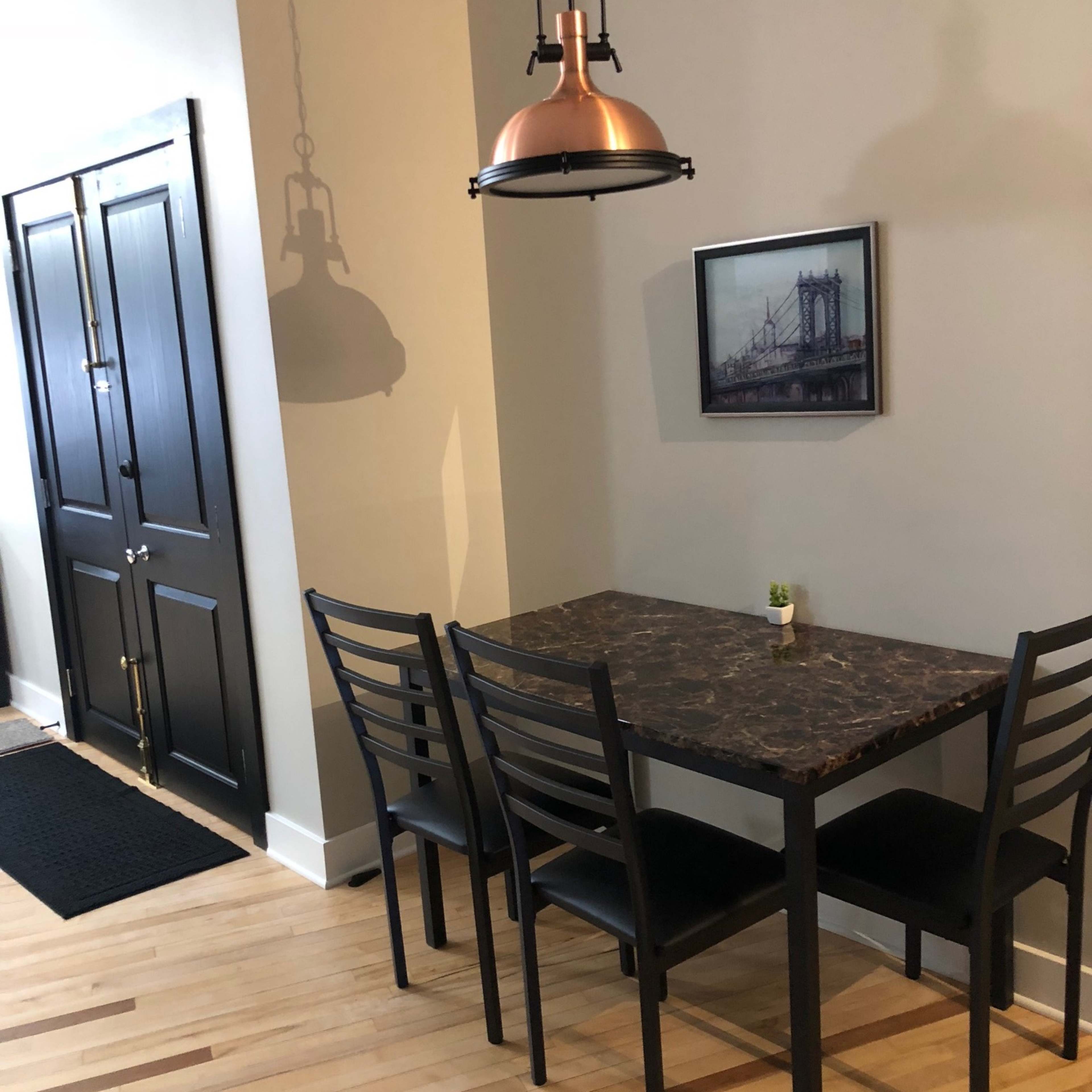 A small dining area features a black table and four chairs beneath a pendant light, with a photo of a bridge on the wall and a doorway to the left.