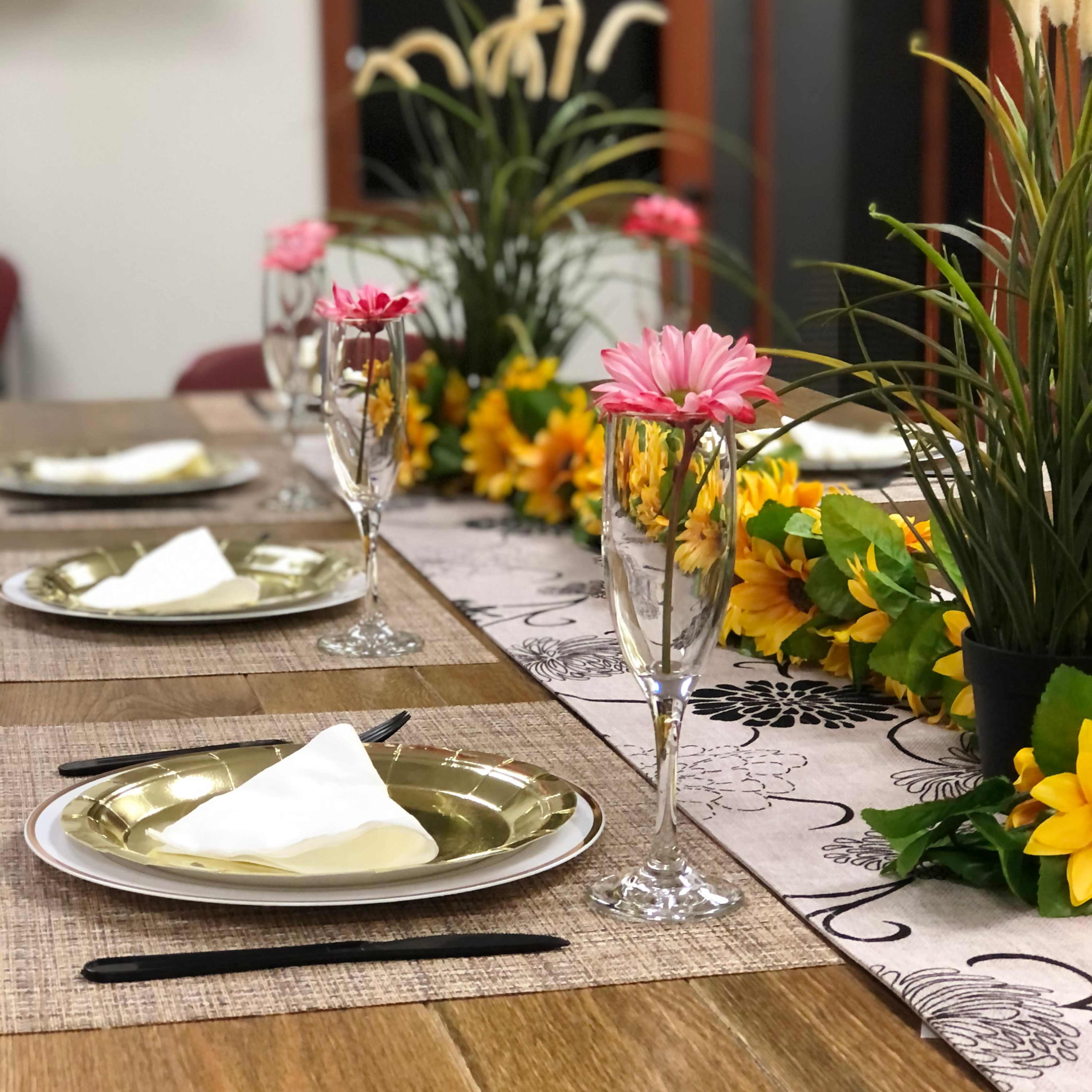 A dining table is set with golden plates, crystal glasses, and floral decorations, including sunflowers and pink flowers.