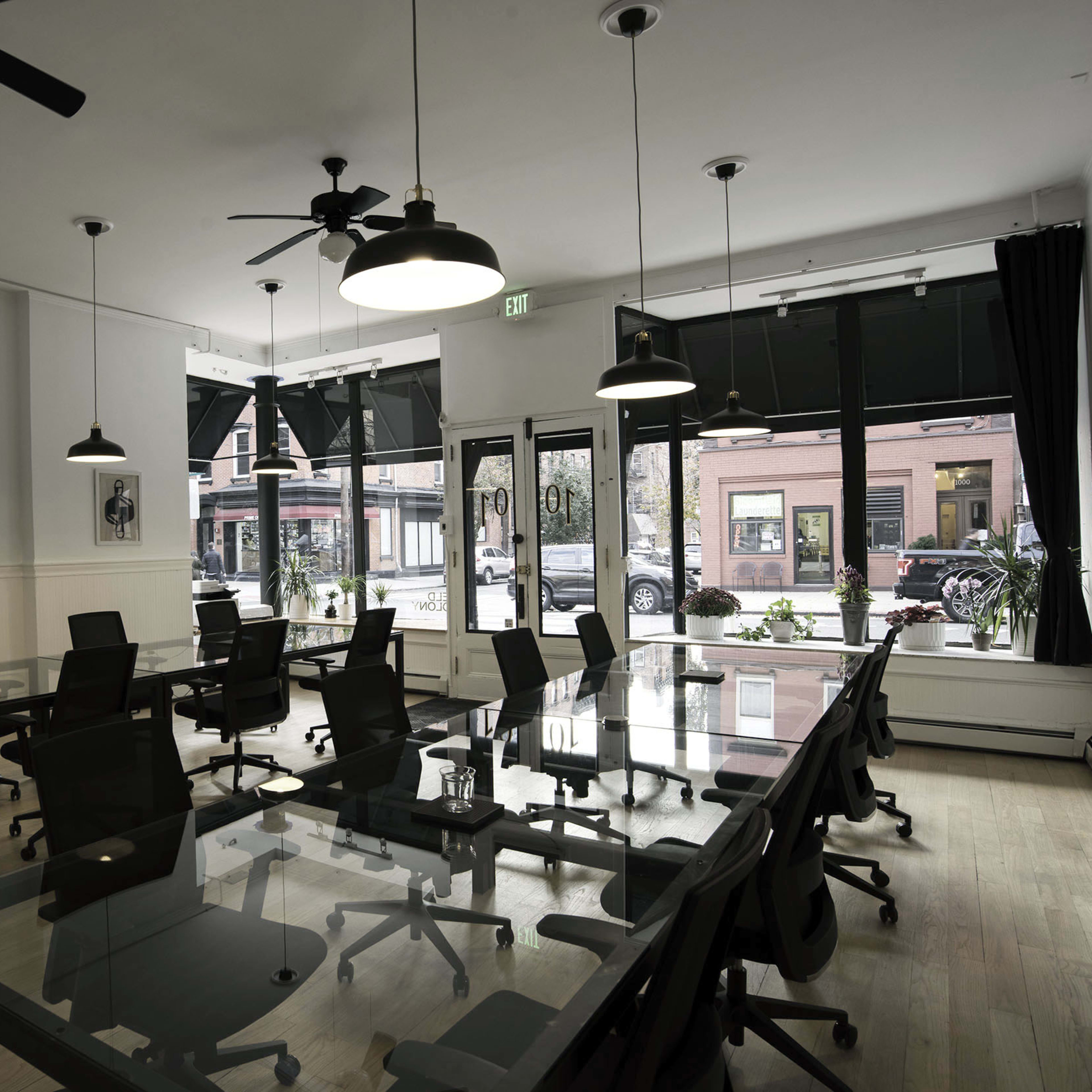 A modern office space with several glass-top tables and black chairs, illuminated by overhead lights and large windows facing an exterior street.