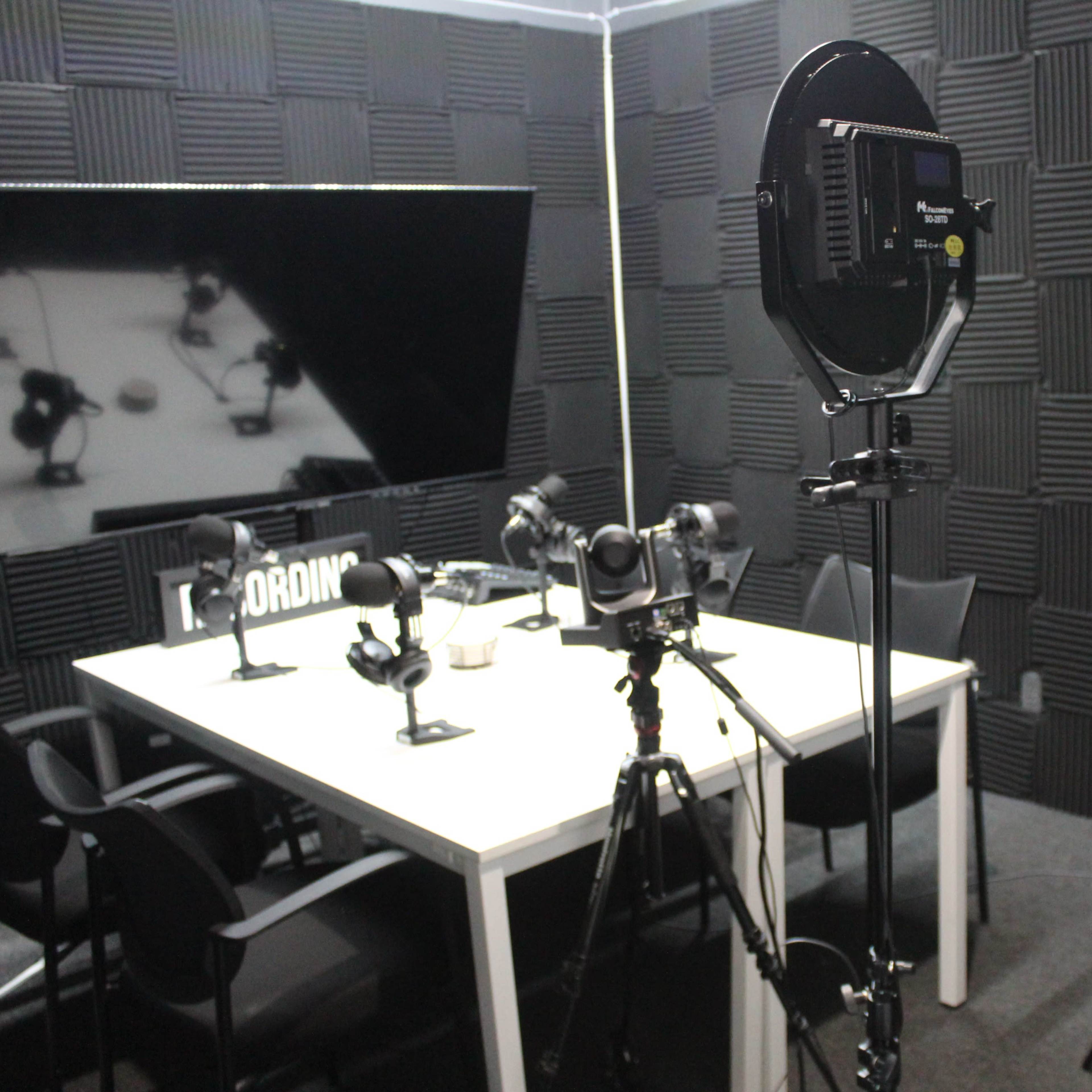 A studio setup with a large screen, multiple cameras, and lighting equipment arranged around a table with microphones.