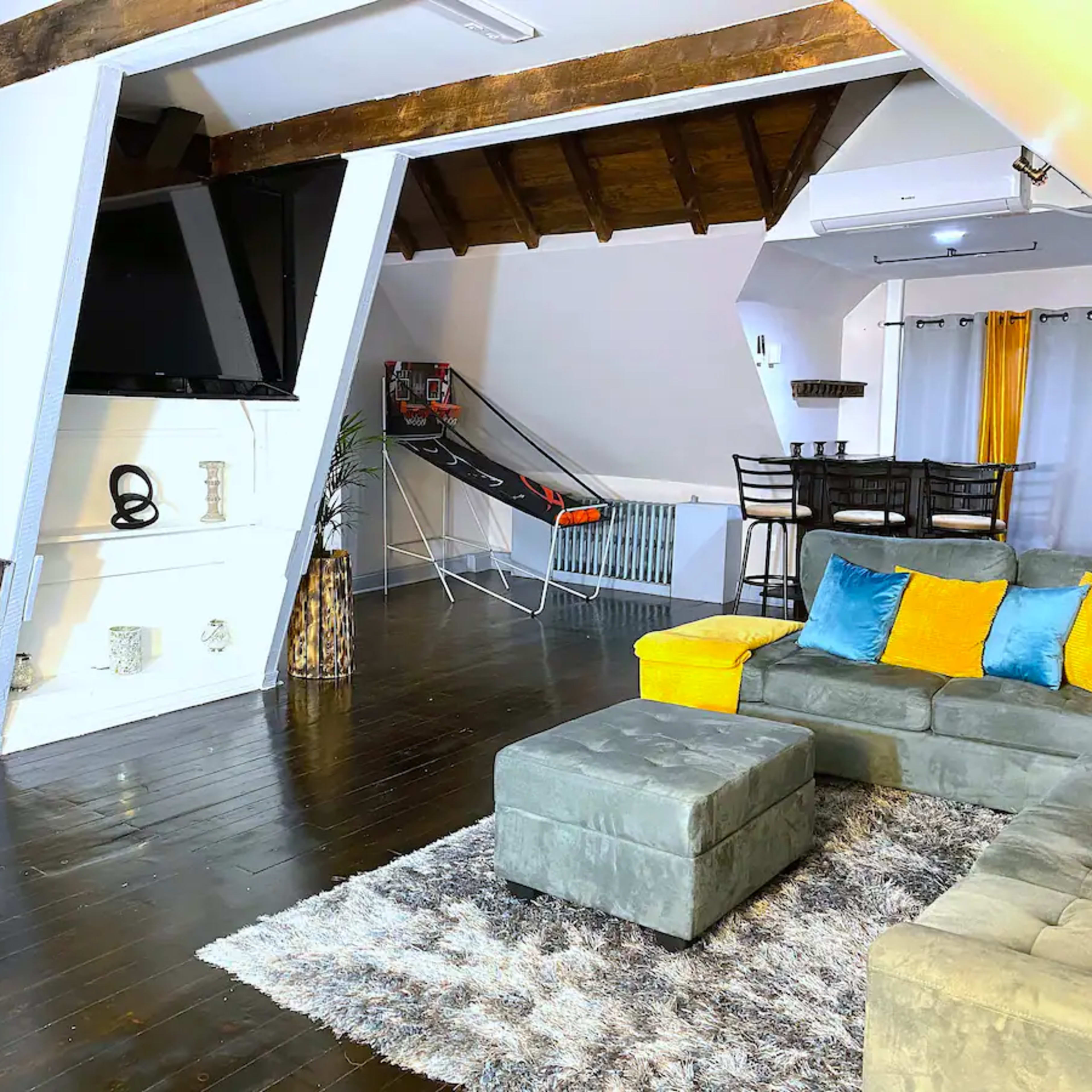 A modern living space with a gray sectional couch, a flat-screen TV mounted on a slanted wall, and a dining area adjacent to it.
