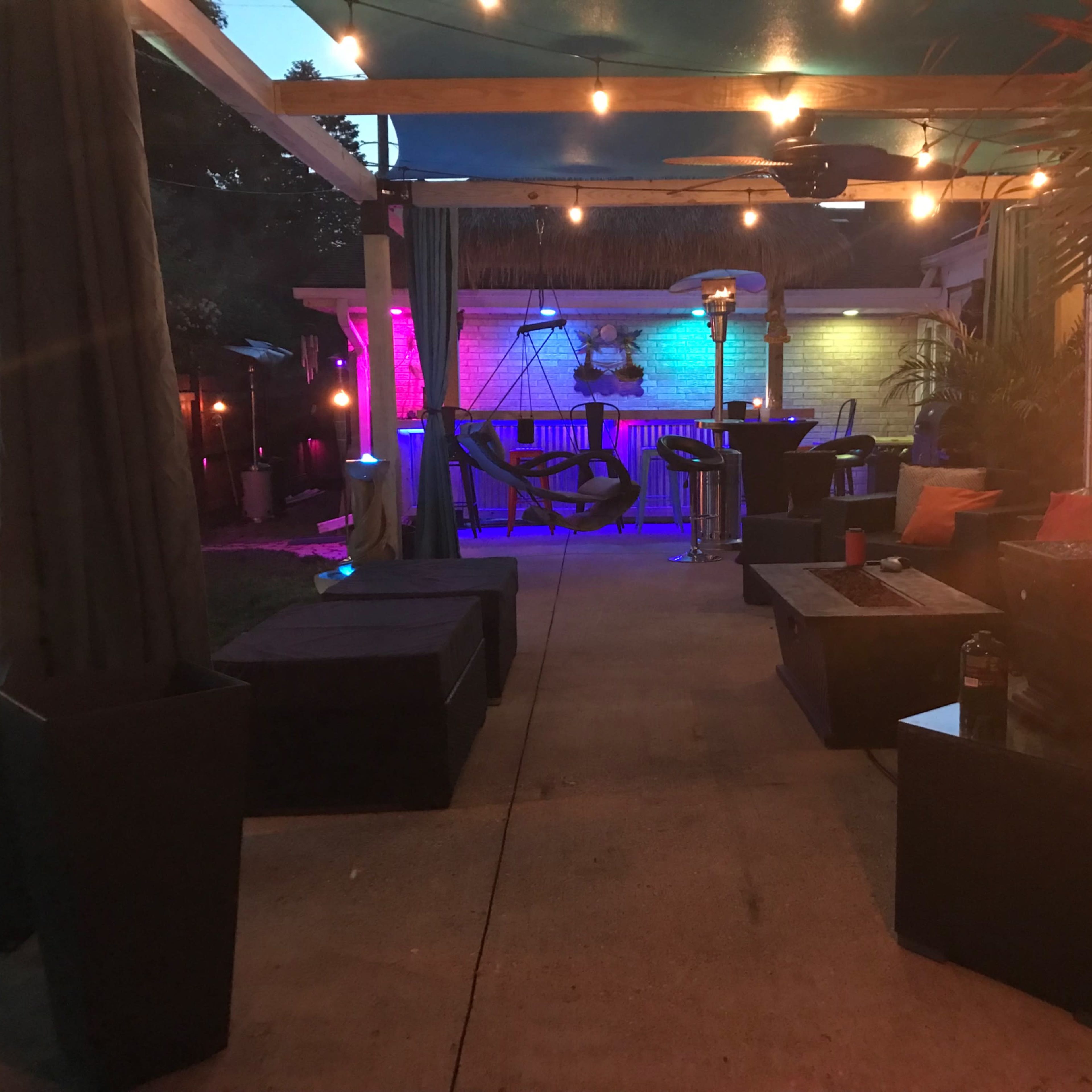 A backyard patio area illuminated by string lights, featuring seating arrangements with cushions, a table, and decorative elements like torches and colorful lighting.