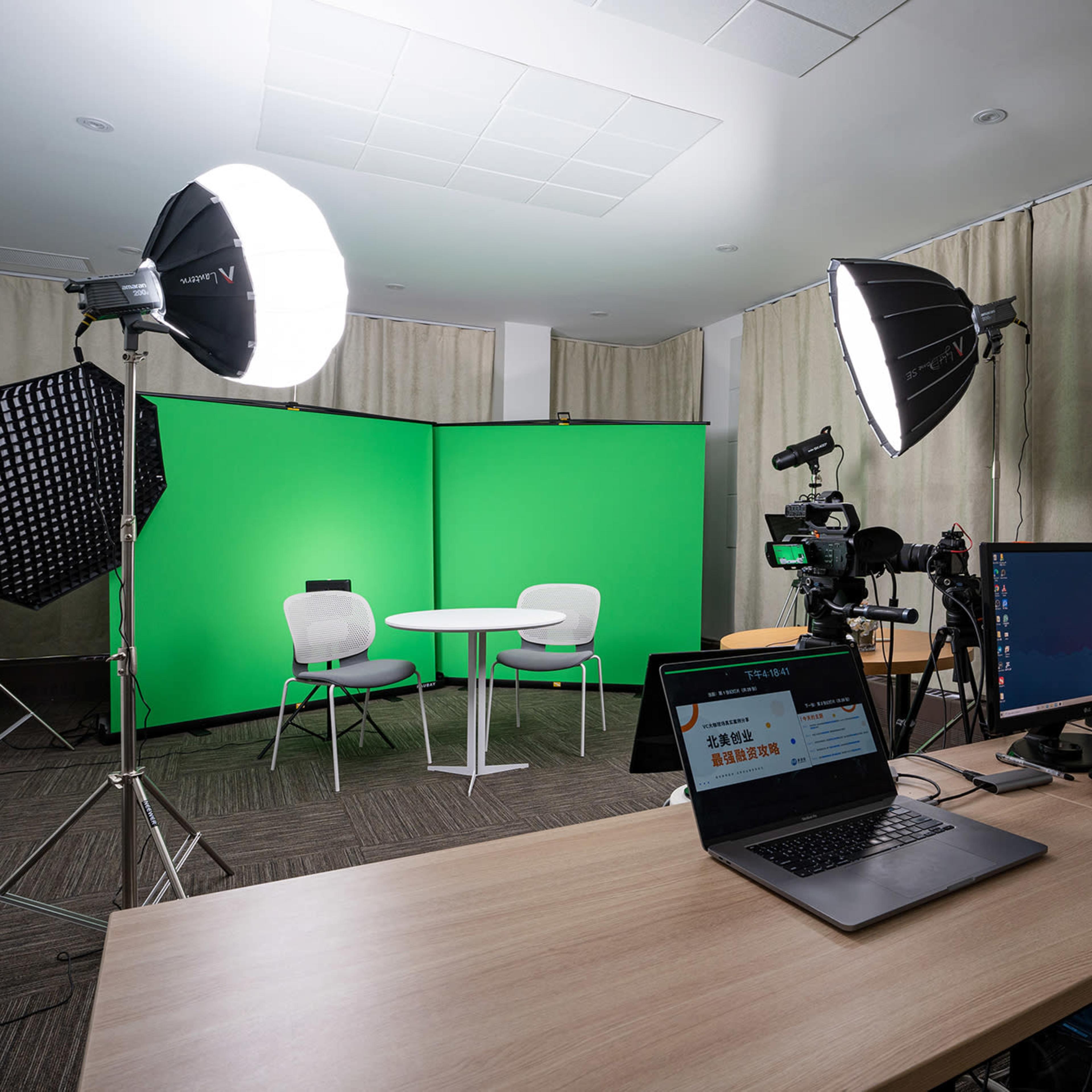 A filming studio features a green screen setup with cameras, lighting equipment, and a table with chairs.
