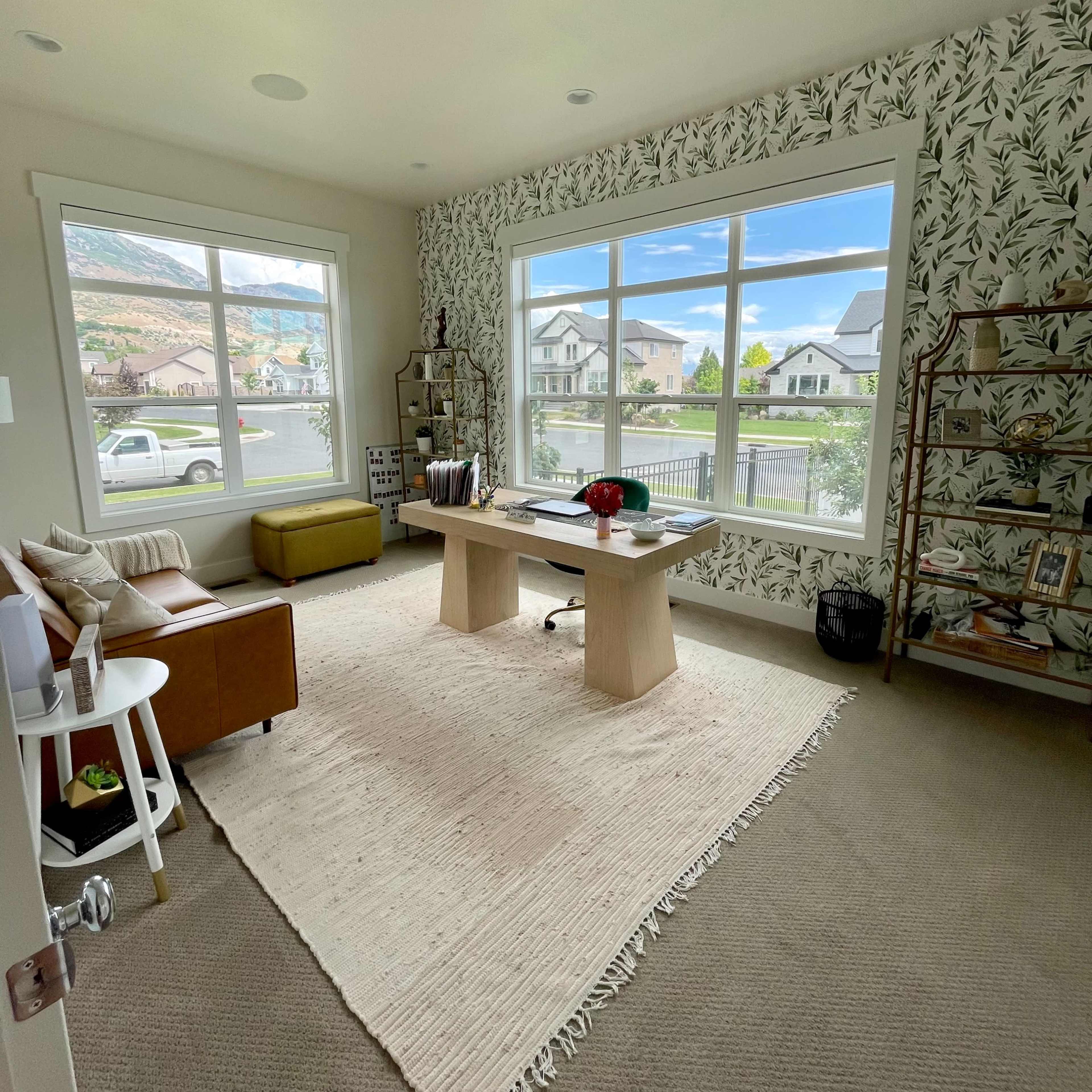 A home office with large windows overlooking a neighborhood, featuring a wooden desk, a rug, and a seating area.