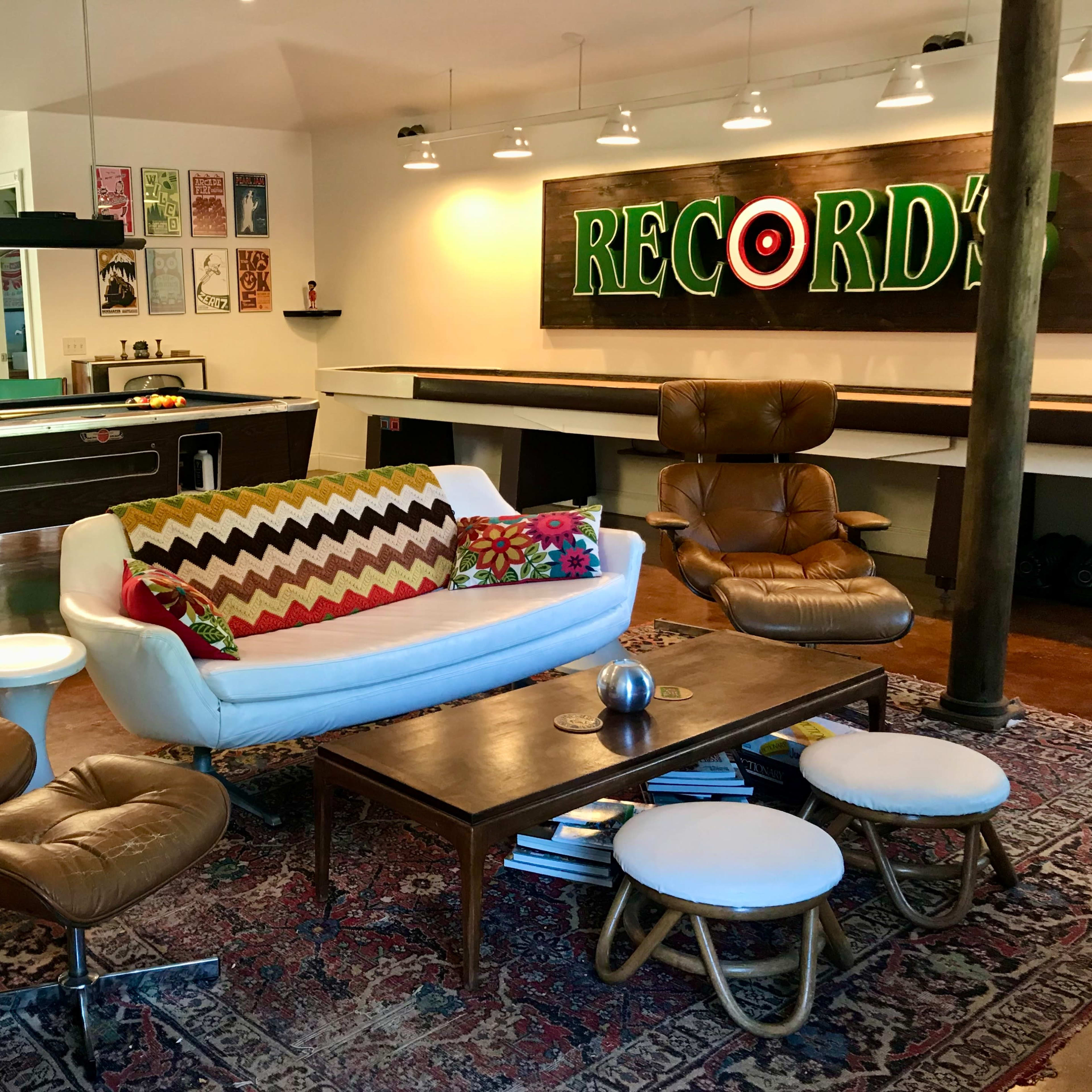 A modern living room with a white sofa, two brown chairs, a wooden coffee table, and a large "RECORD'S" neon sign on the wall.