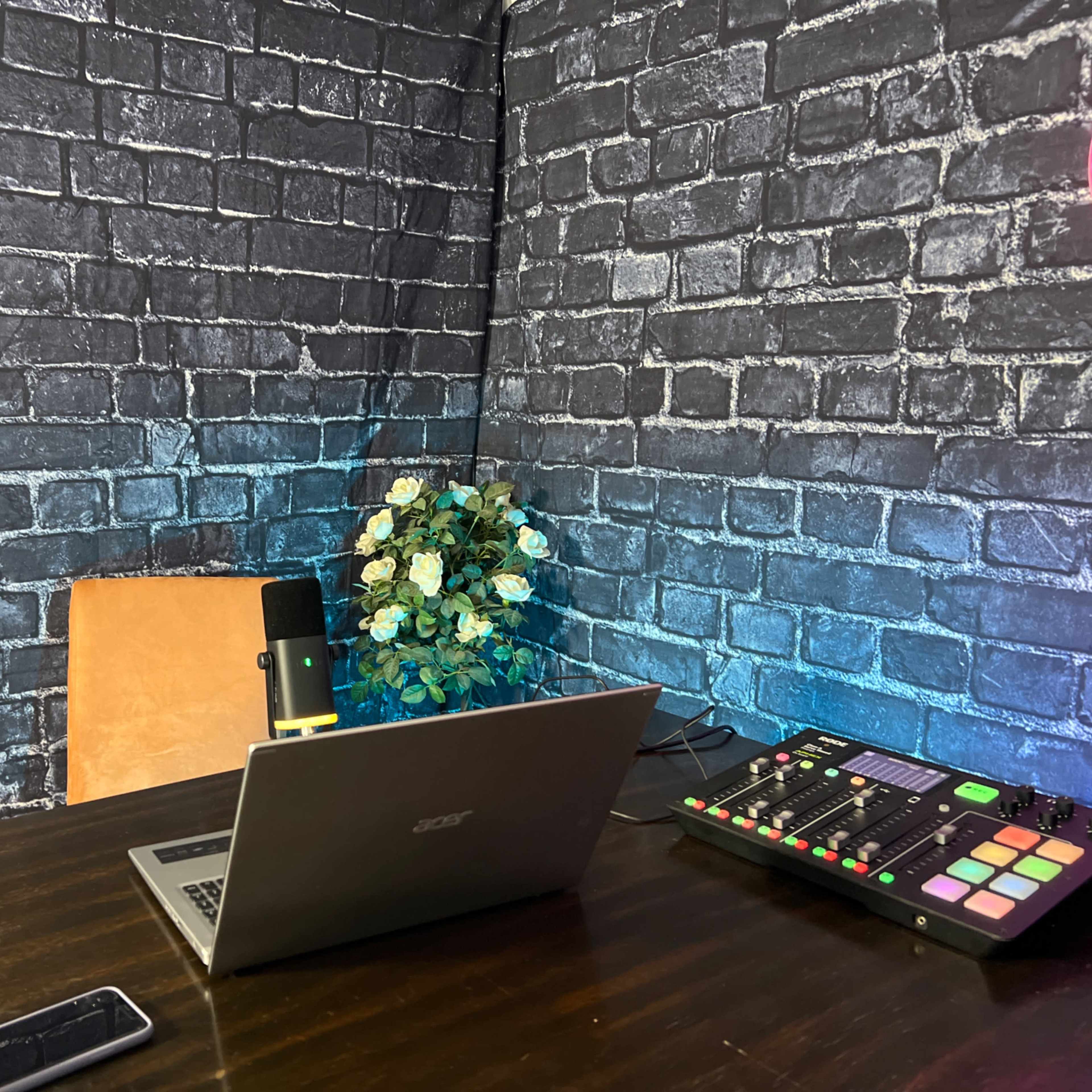 A wooden table with a laptop, microphone, and music mixer is set against a backdrop of black brick walls and a small plant.